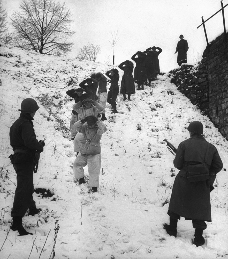 German prisoners, some of them wearing coveralls for camouflage in the snow, are herded by guards. (In close fighting, U.S. troops also used snow-camouflage suits.)
