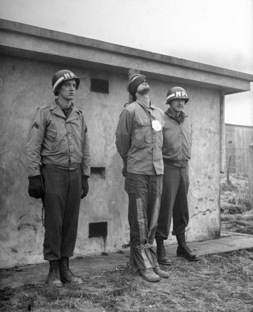 A blindfolded prisoner is securely bound, hand and foot, to a stake in front of a concrete wall. A large white paper target is pinned over his heart. American MPs stand at attention until the firing squad's commanding officer inspects the final arrangements. Belgium, 1944.
