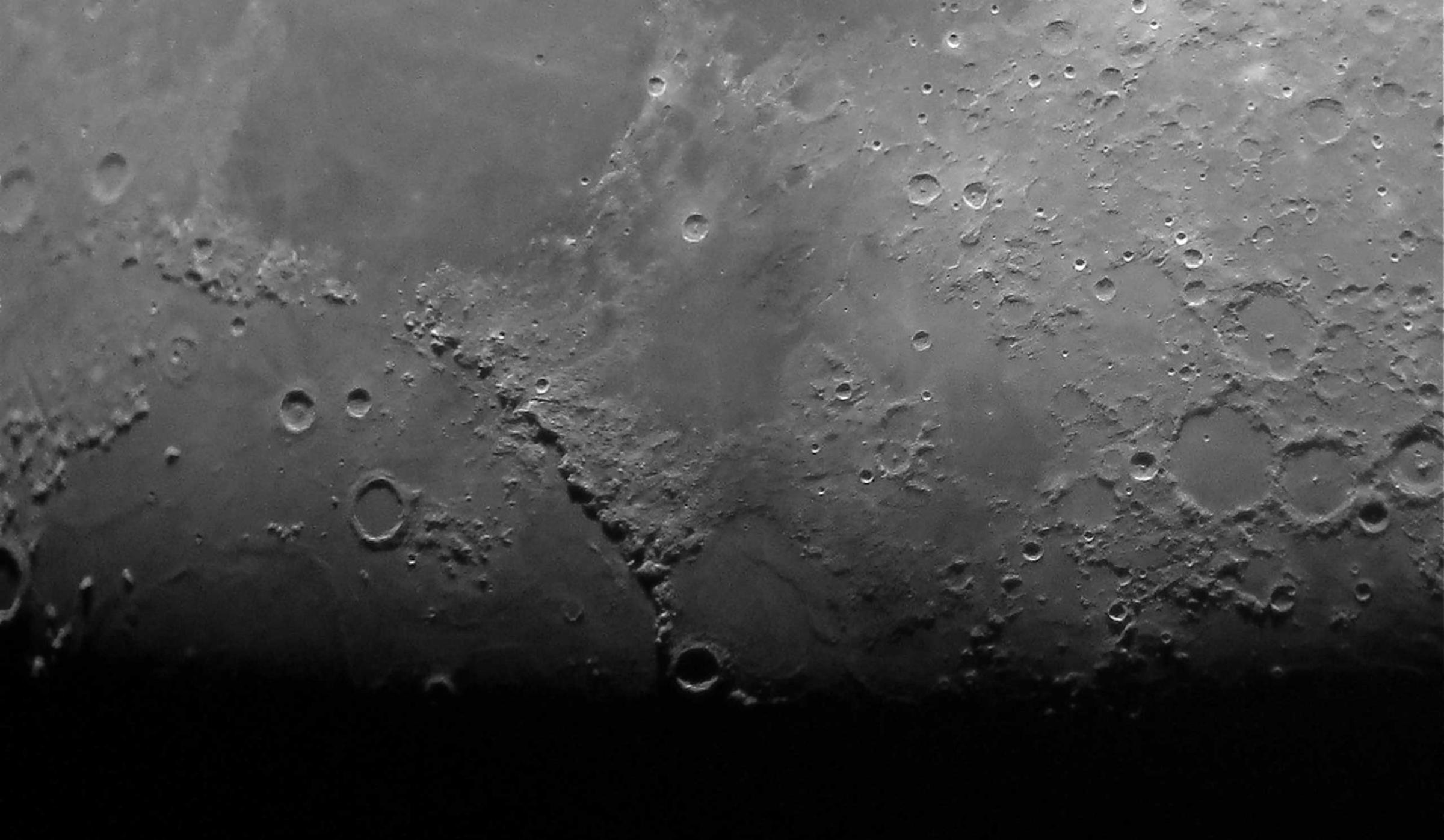 A beautifully sharp and artfully framed detail of the Moon. The terminator which separates the daytime and night-time parts of the Moon is aligned with the bottom edge of the photograph. The Sun's light shines at a low angle onto the surface of the Moon just above this line, showing the contrast between smooth maria and rugged crater rims to the best advantage.