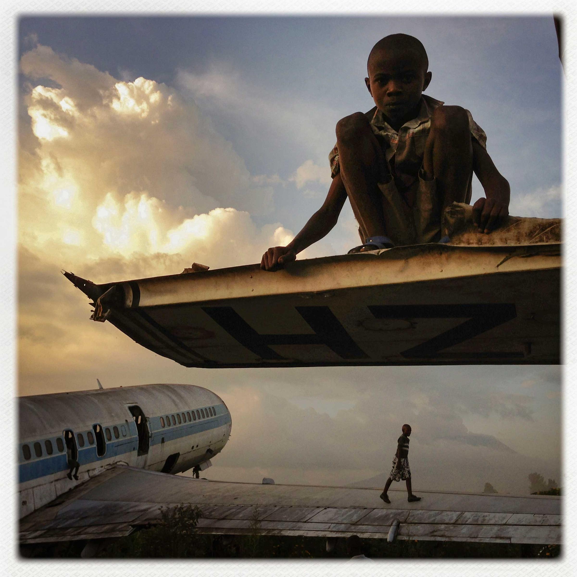 CONGO. Goma. December 14, 2012. Abandoned planes are a common site at airports in Africa. At Goma Airport, in the Democratic Republic of Congo, planes left due to wars and volcanic eruptions over the past two decades have become a playground for street children, some of whom sell the parts which are made into stoves and other items to be sold on the streets of Goma. One is generally prohibited from photographing this airport but in mid-December, 2012, after the M23 rebel force which occupied Goma left and before the FARDC (military of the D.R.C.) returned to the city, a security vacuum meant that nobody was guarding this section of the airport. Children guided me through the planes, which were later discussed by my Congolese fixer:‚ÄúIn January of 2002, the volcano (Nyiragongo, just outside Goma) exploded and the lava blocked the planes. I helped move this plane after I and many of my friends living near the airport lost our homes to lava, on the first day of the eruption. On the second day, we saw the lava moving towards the planes. I and others were just watching the lava flow getting closer to the planes and we decided to move one of them, this newer one. There were at least a hundred people there pushing the plane for about 300 meters. A friend mine, who was there and whose house was also destroyed, had a childhood dream to be a pilot. But his parents were too poor and all the schools were expensive, so he could not hold onto that dream. He forgot about it, but then on that day, when we needed to move the plane, he told me to help him inside so he might steer it! We all pushed the plane as my friend waved his arm out the window, in the cockpit. We then climbed in the plane and saw the lava flowing down the volcano and into town.‚Äù