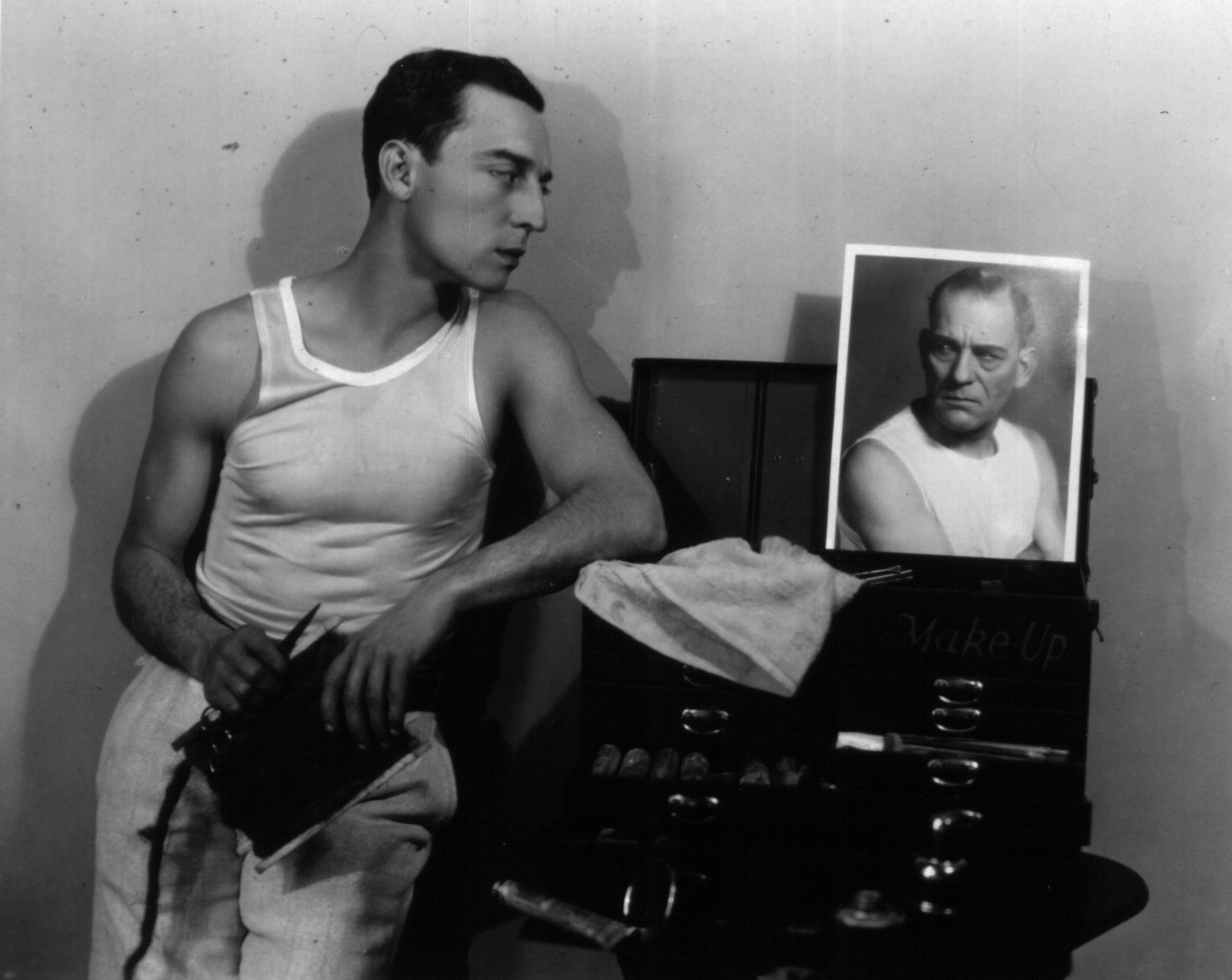 American comic actor Buster Keaton engages in a staring match with a photograph of Lon Chaney as he prepares to apply his make-up.  1927
