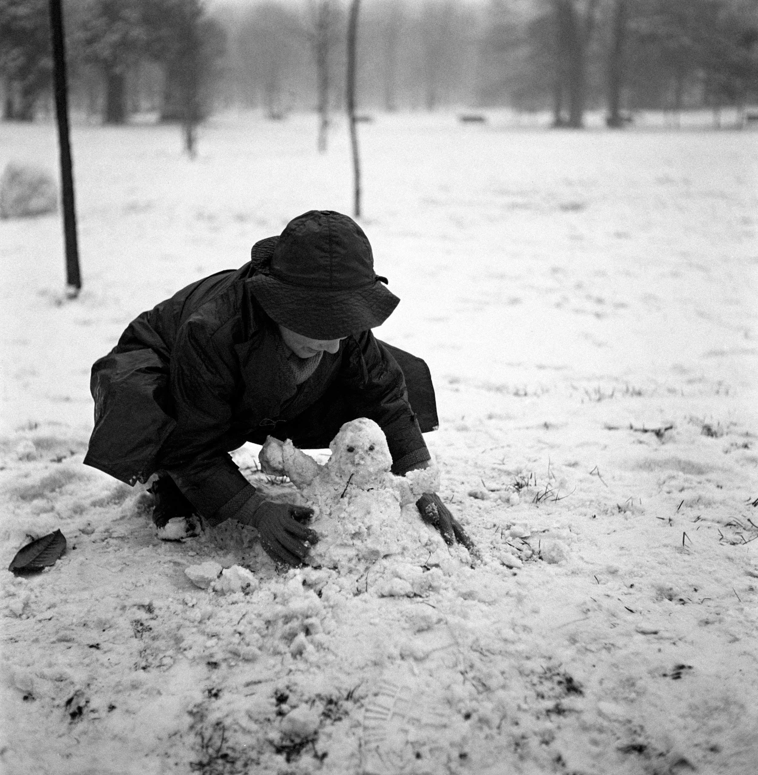 A little boy crouching and making a snowman in a park after a snowfall in Milan in the 1950s.