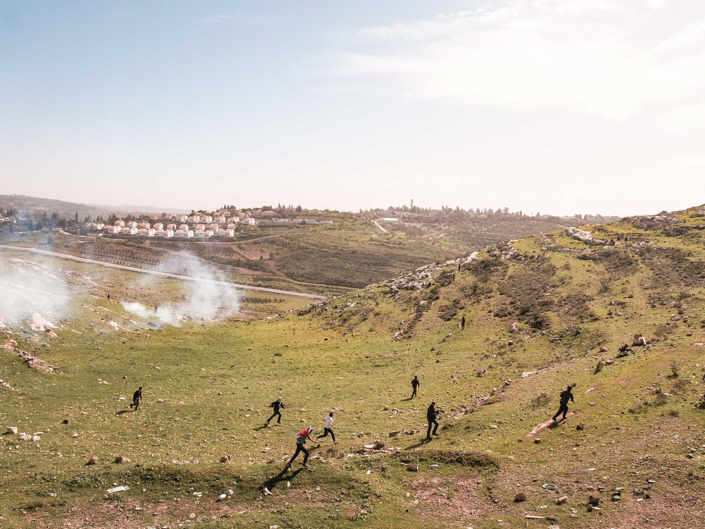 West Bank, Nabi Saleh. 2013. Protestors run away as Israeli soldiers fire tear gas at them. The Israeli settlement of Halimish is in the background.Residents of Nabi Saleh have been protesting the Israeli Occupation every Friday after midday prayers since 2009. The protests started after Israelis from the nearby settlement of Halimish took over a small spring that had been on Palestinian land. After widening and adding a bench to the spring, the settlers refused to allow the Palestinians to continue using it. The weekly protests quickly devolve into rock throwing by the Palestinians and tear gas, rubber bullets, and 'skunk' (a spray mixed with water that has a horrific stench that can linger for weeks) in response by the Israelis. (Peter van Agtmael / Magnum Photos)