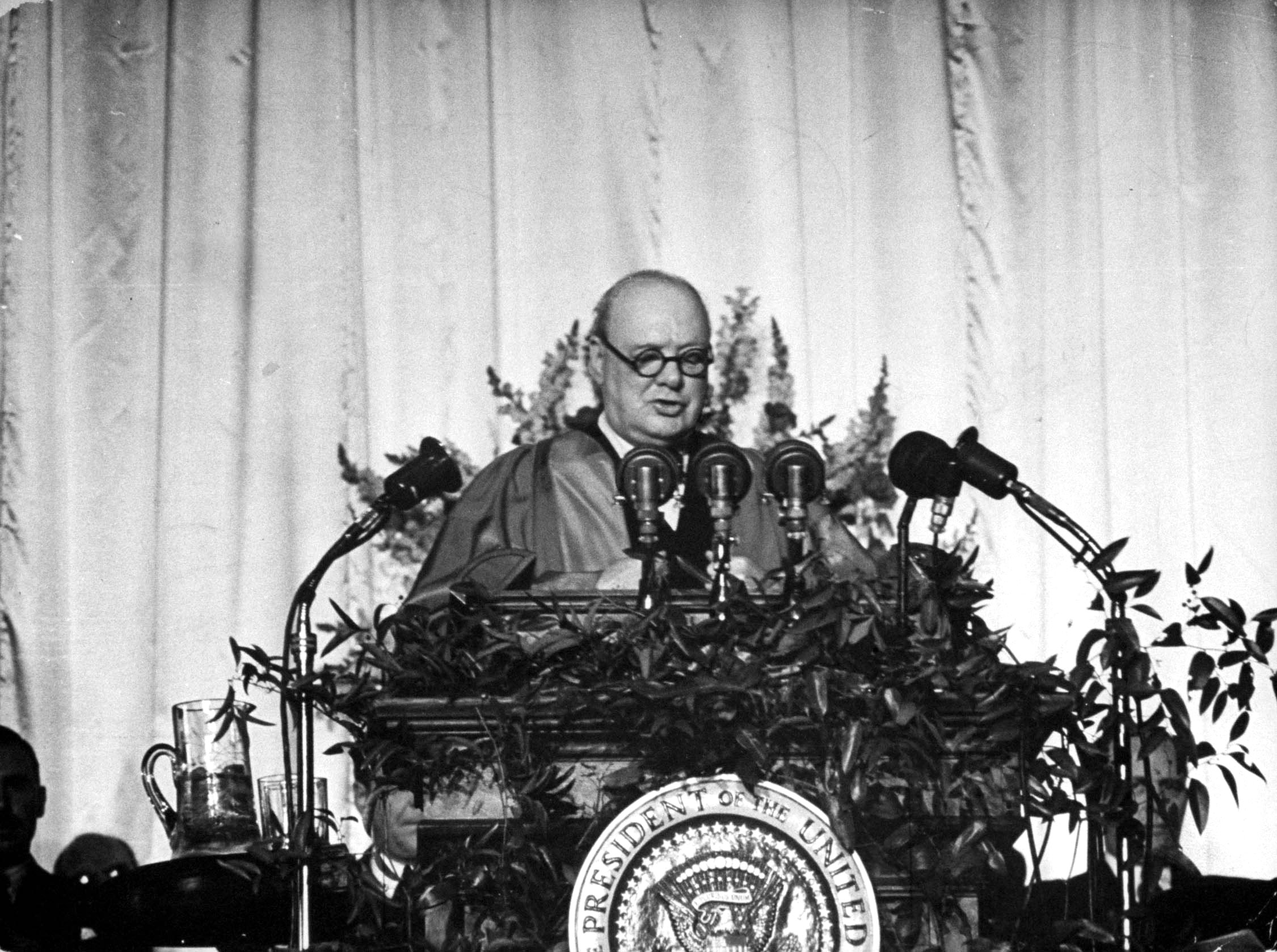 Churchill speaks in the gym from a rostrum decorated with shrubs and radio microphones. His small audience sat on hard, borrowed bleachers.