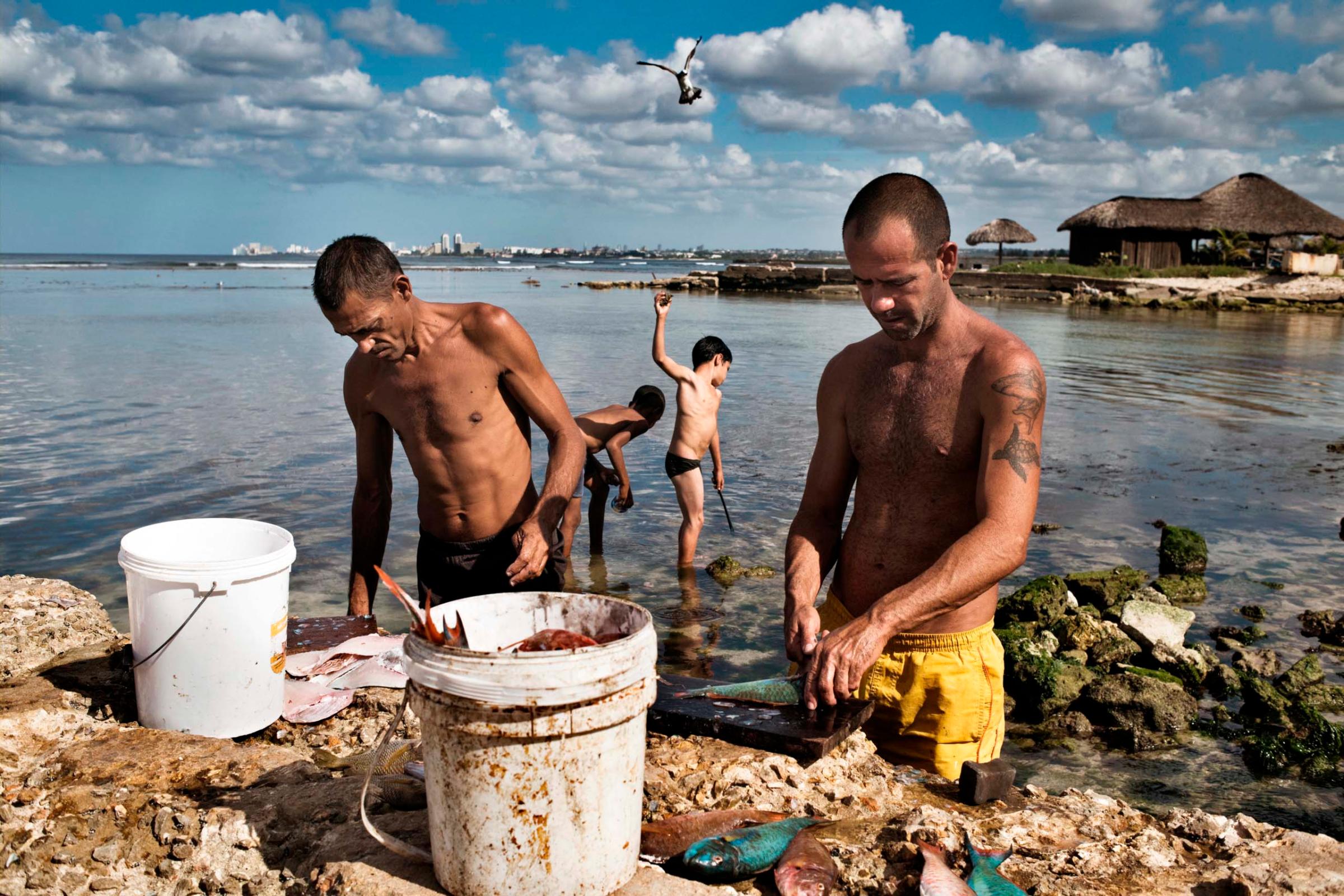 January 2015. Men clean fish, while children play by the shore, in the Jaimanitas neighborhood of Havana.