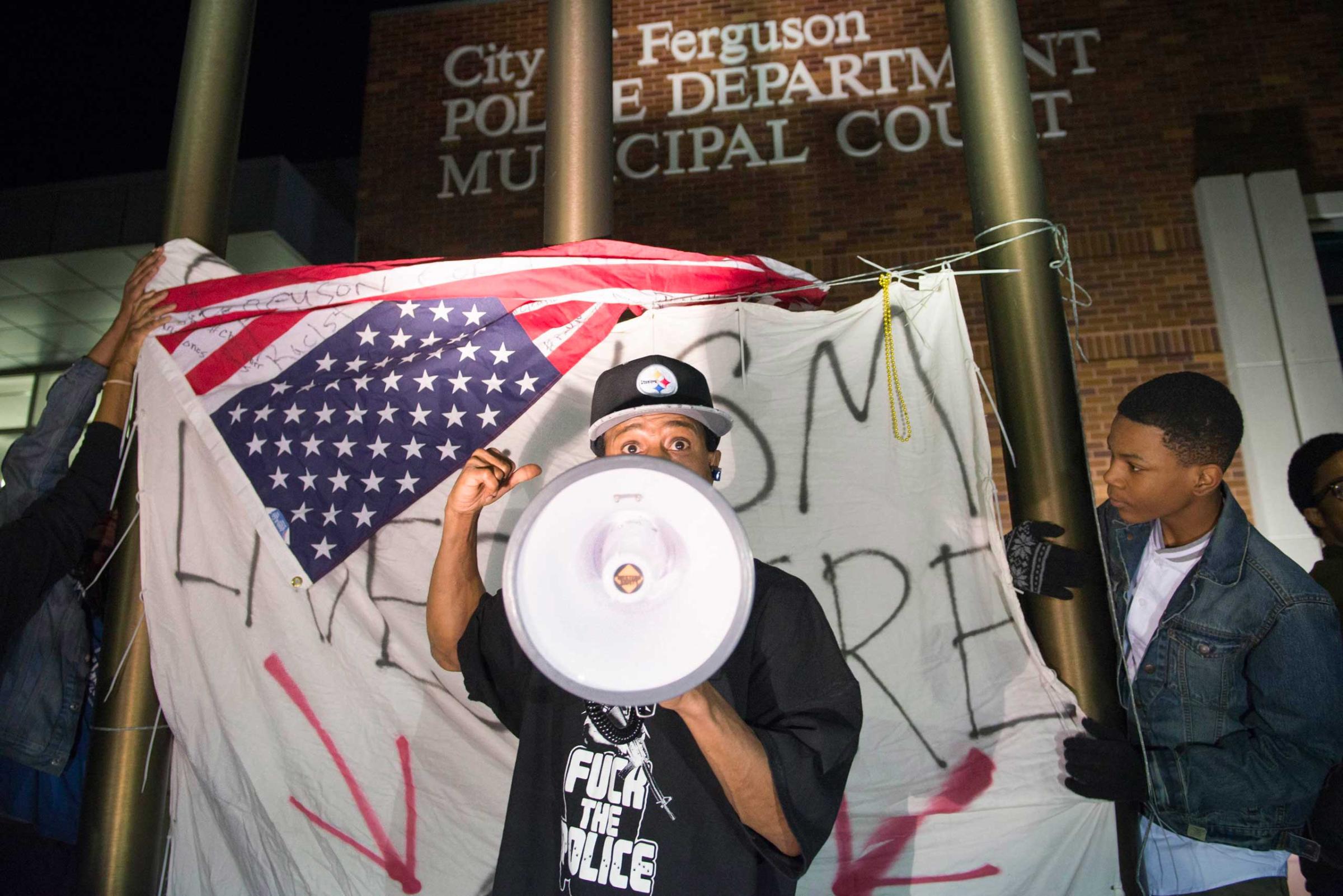 A protester chants in front of a flag which reads, "Racism lives here", outside the City of Ferguson Police Department and Municipal Court in Ferguson, March 11, 2015.