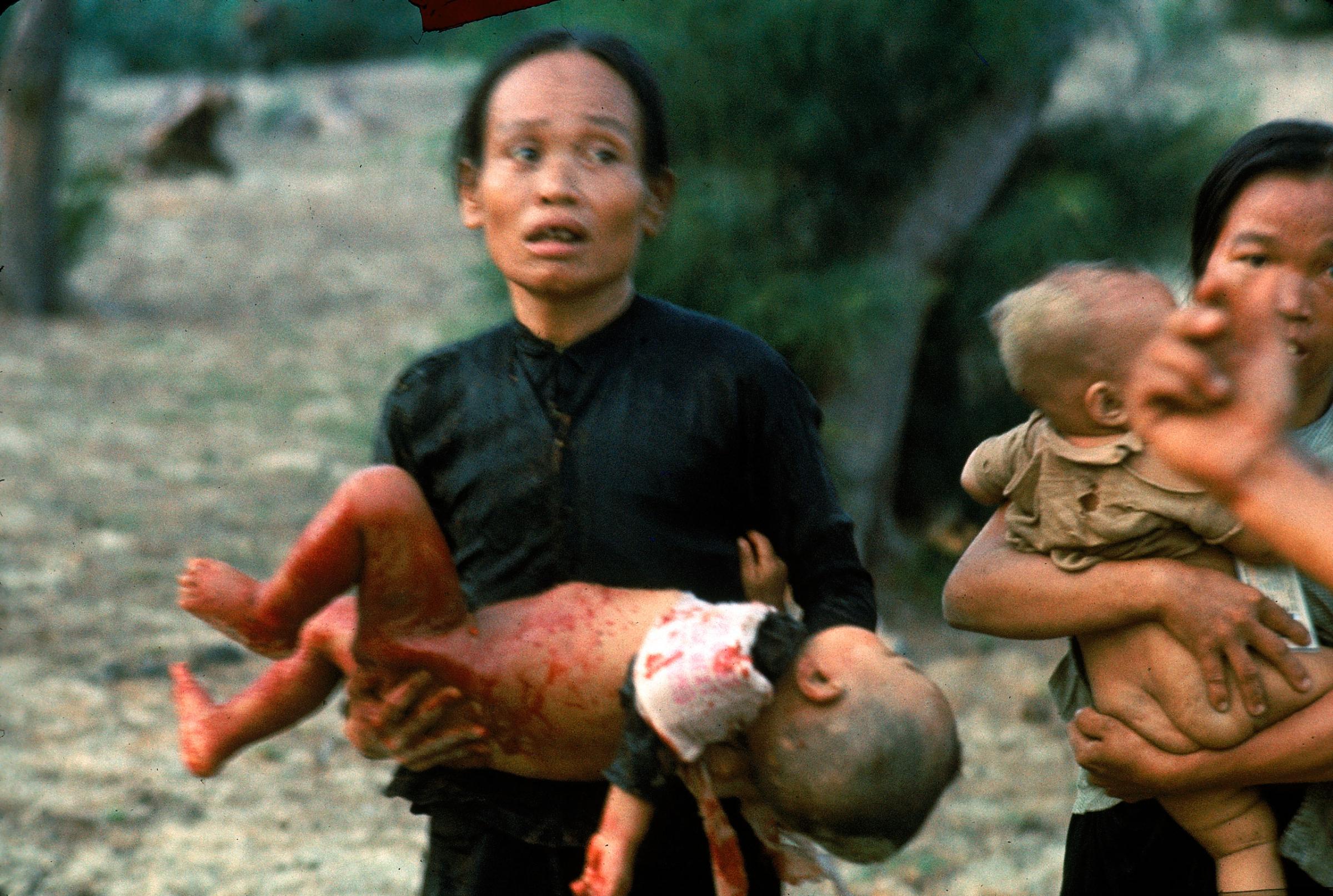 Screaming with terror amid the furious noises of war, a Vietnamese woman clutches her blood-drenched child who was wounded when jets strafed before landing. Before the air strikes, loudspeaker helicopters broadcast warnings, but many villagers were too panicked to follow instructions and evacuate. Distraught, this woman blundered into the thick of the battle.