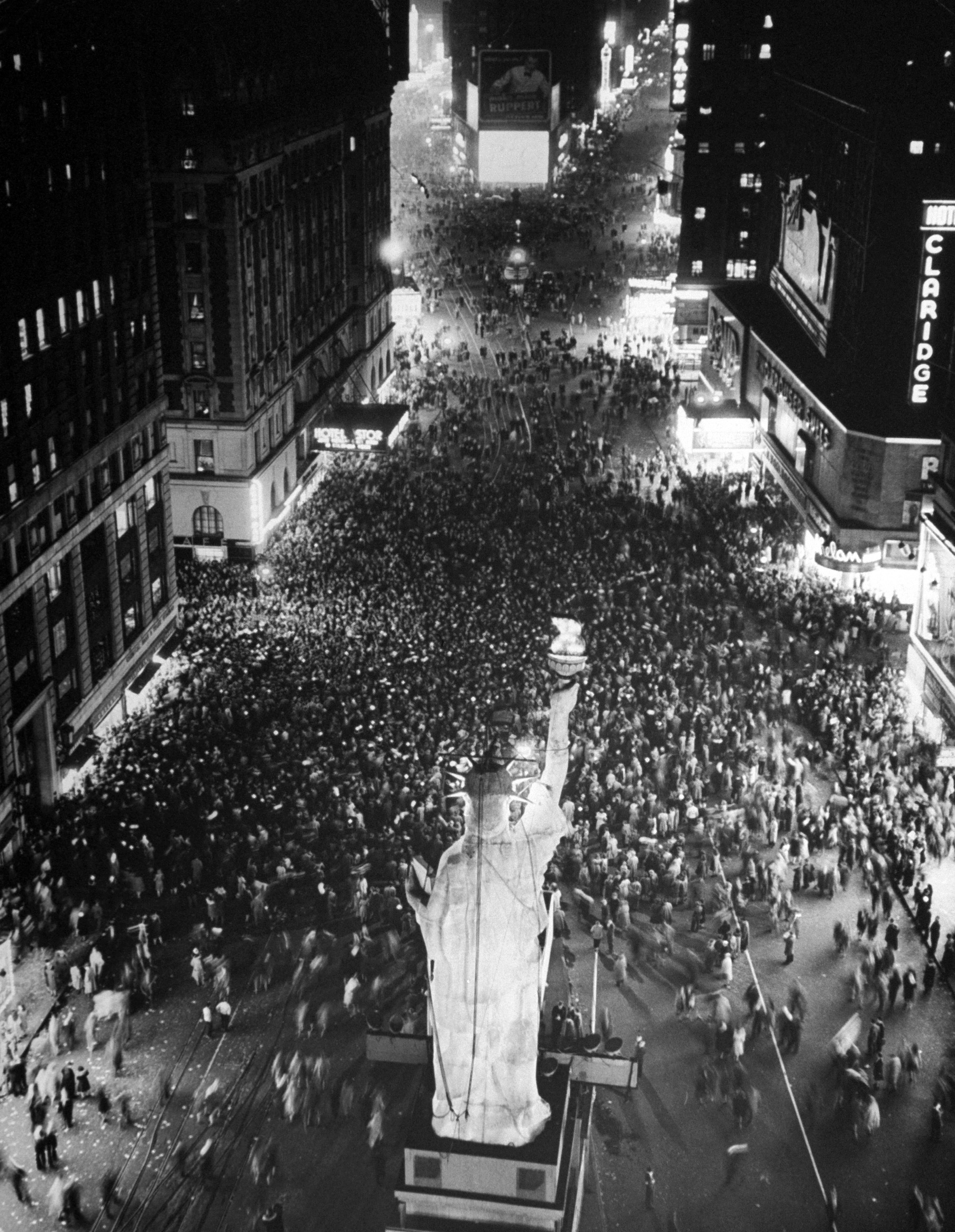Night time crowd of people in Times Square celebrating end of the War in Europe, VE Day, in front of lighted replica of the Staue of Liberty.