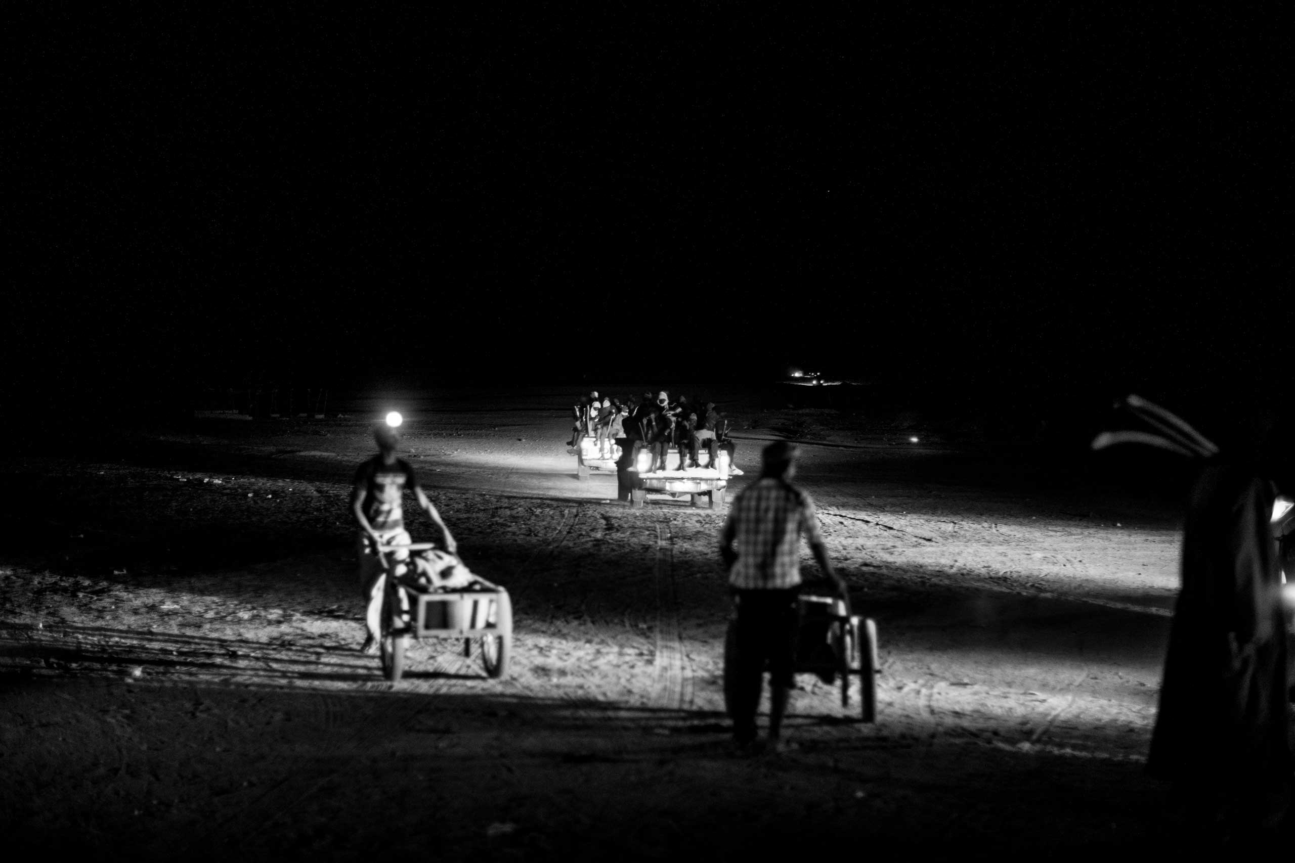 Trucks carrying migrants pass back into the desert as vendors push their carts back to wait for more trucks at a small desert outpost a few hours north of Agadez. Smugglers time their departure into the desert to within a few hours, forming a loose caravan in the hopes that it will provide some shred of security against the increasingly lawless Sahara.
