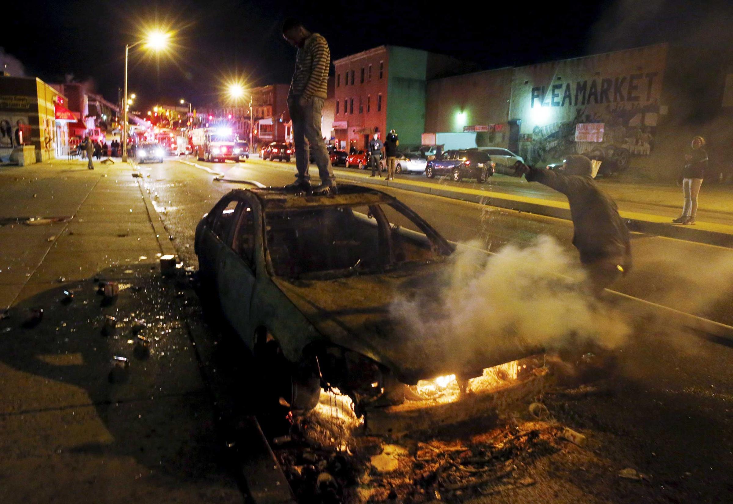 A rioter stands atop a burning car as another man pours fuel onto the fire while Baltimore firefighters behind them fight fires in mutliple burning buildings set ablaze by rioters during clashes in Baltimore