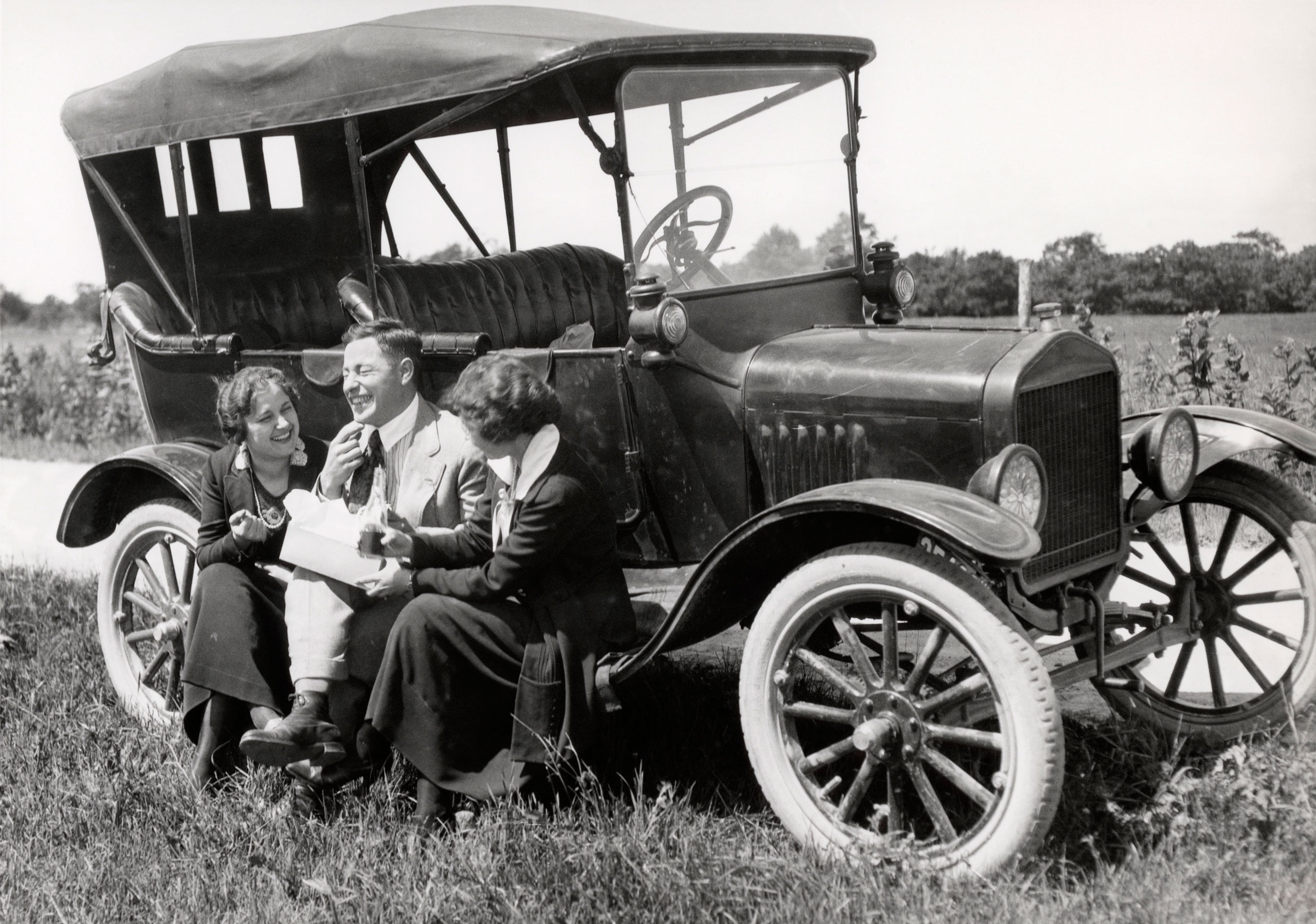 Two women and one man sitting on the running board of a Model T Ford eating lunch, 1920.