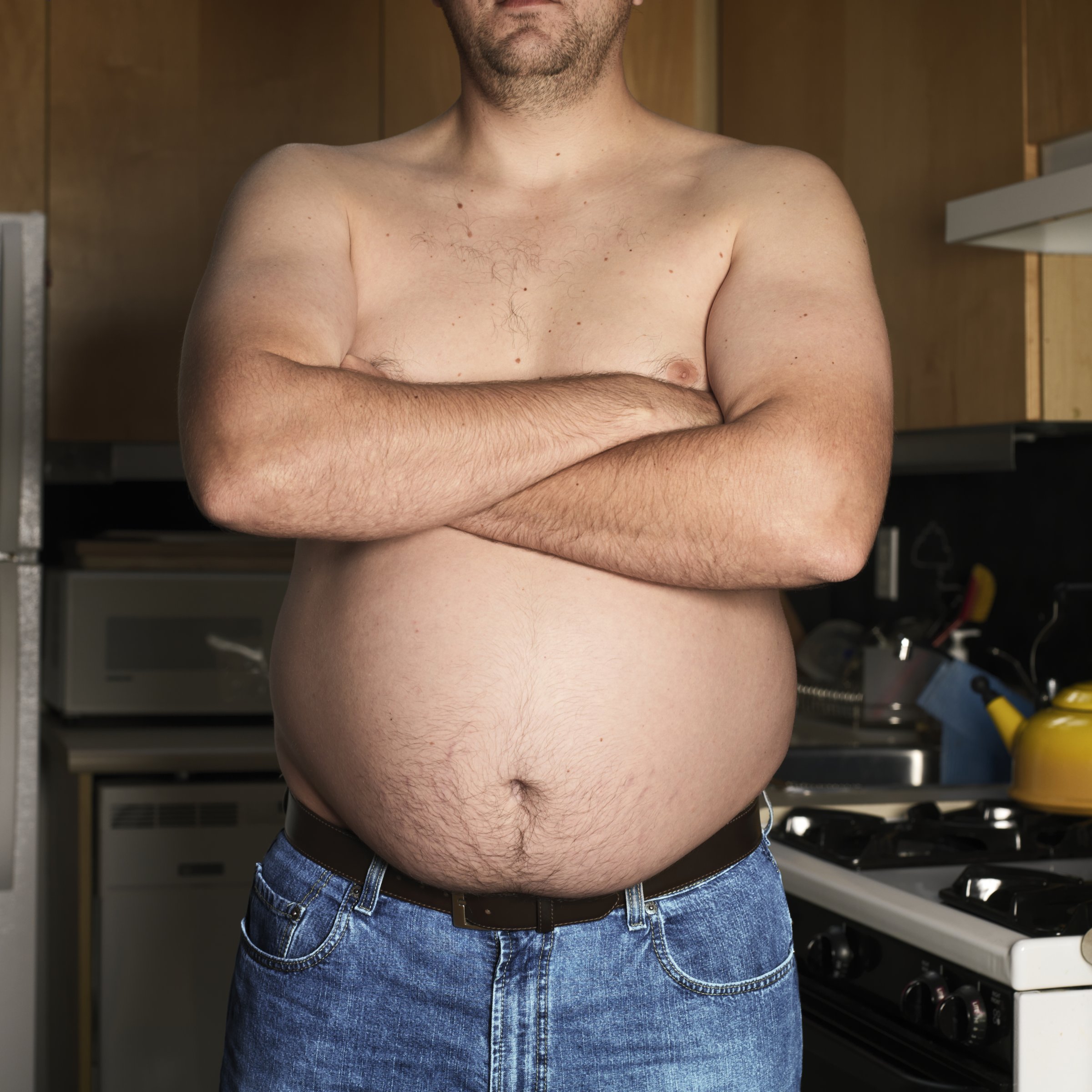 Shirtless man standing in kitchen with arms crossed, mid section, close-up