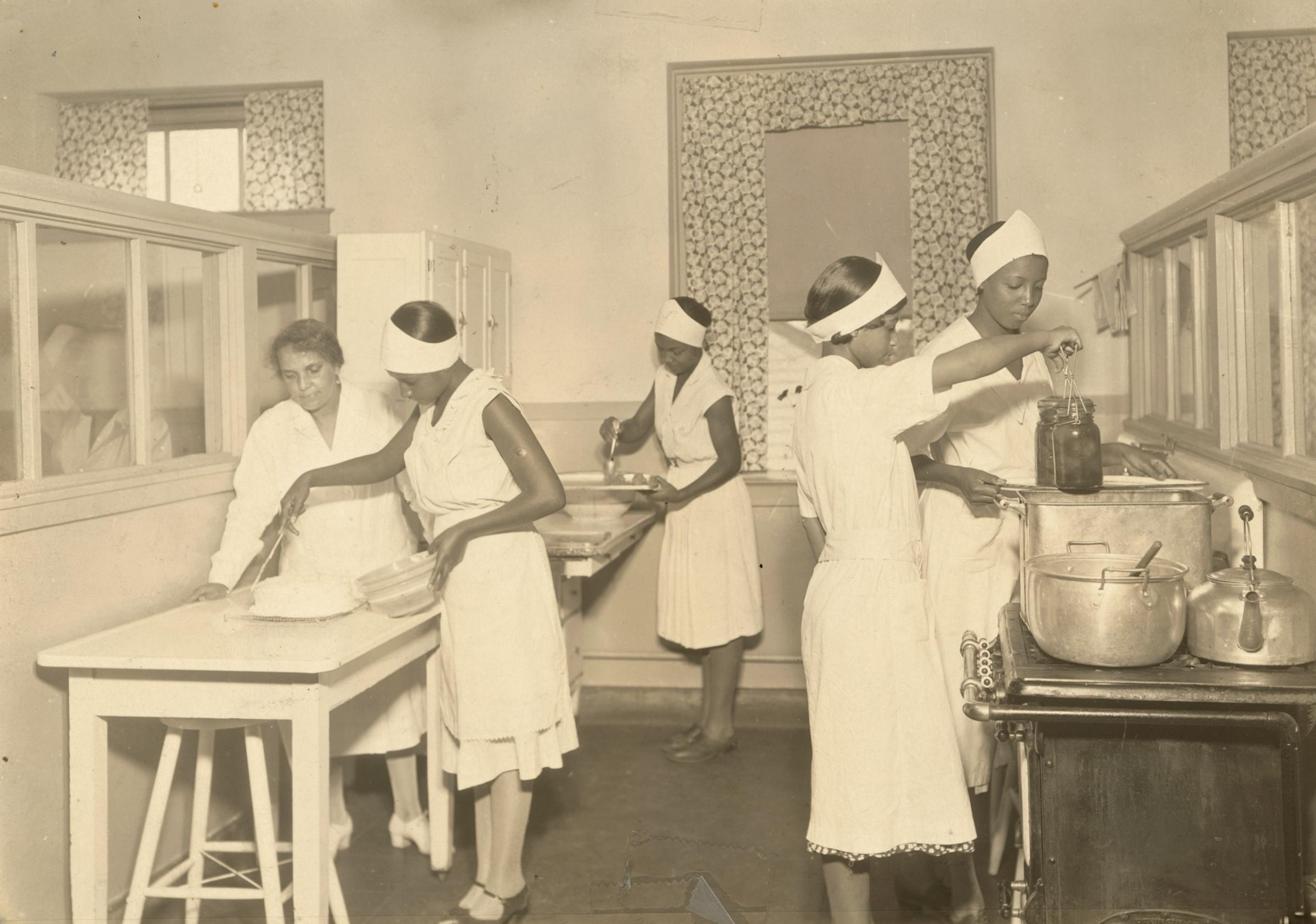 Four women working in a kitchen at the Manual Training and Industrial School for Colored Youth in Bordentown, NJ. A fifth, older woman oversees a younger as she ices a cake, 1935.