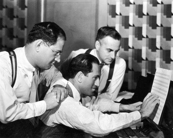 Circa 1925: George Gershwin (1888 - 1937) (C) at work as his brother and songwriting partner, lyricist Ira Gershwin (1896 -1983) (L), and British dramatist Guy Bolton (1884 - 1979), look on.