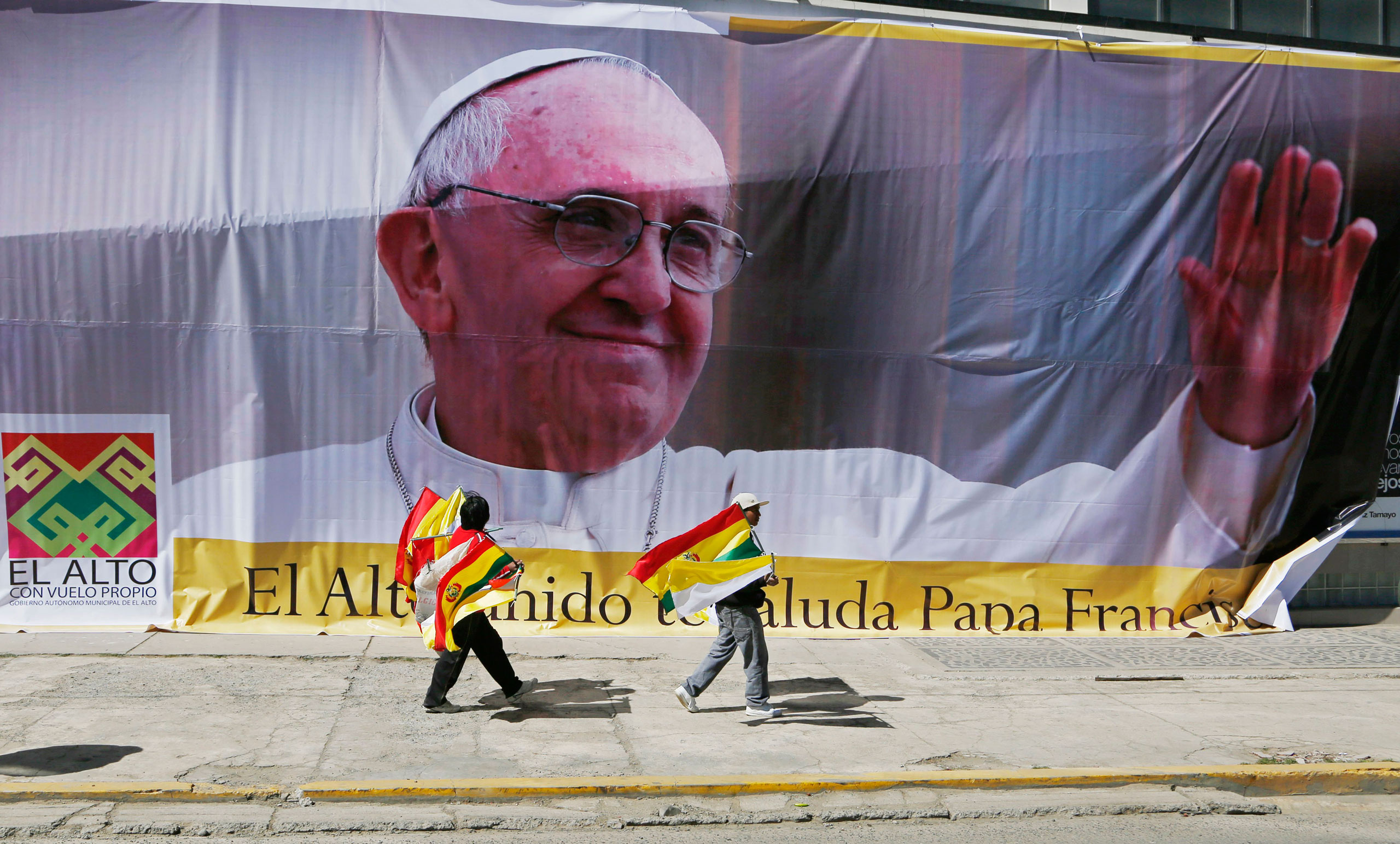 Street vendors selling Bolivian and Vatican flags pass a large image of Pope Francis ahead of the Pope's arrival to El Alto, Bolivia, on July 8, 2015.