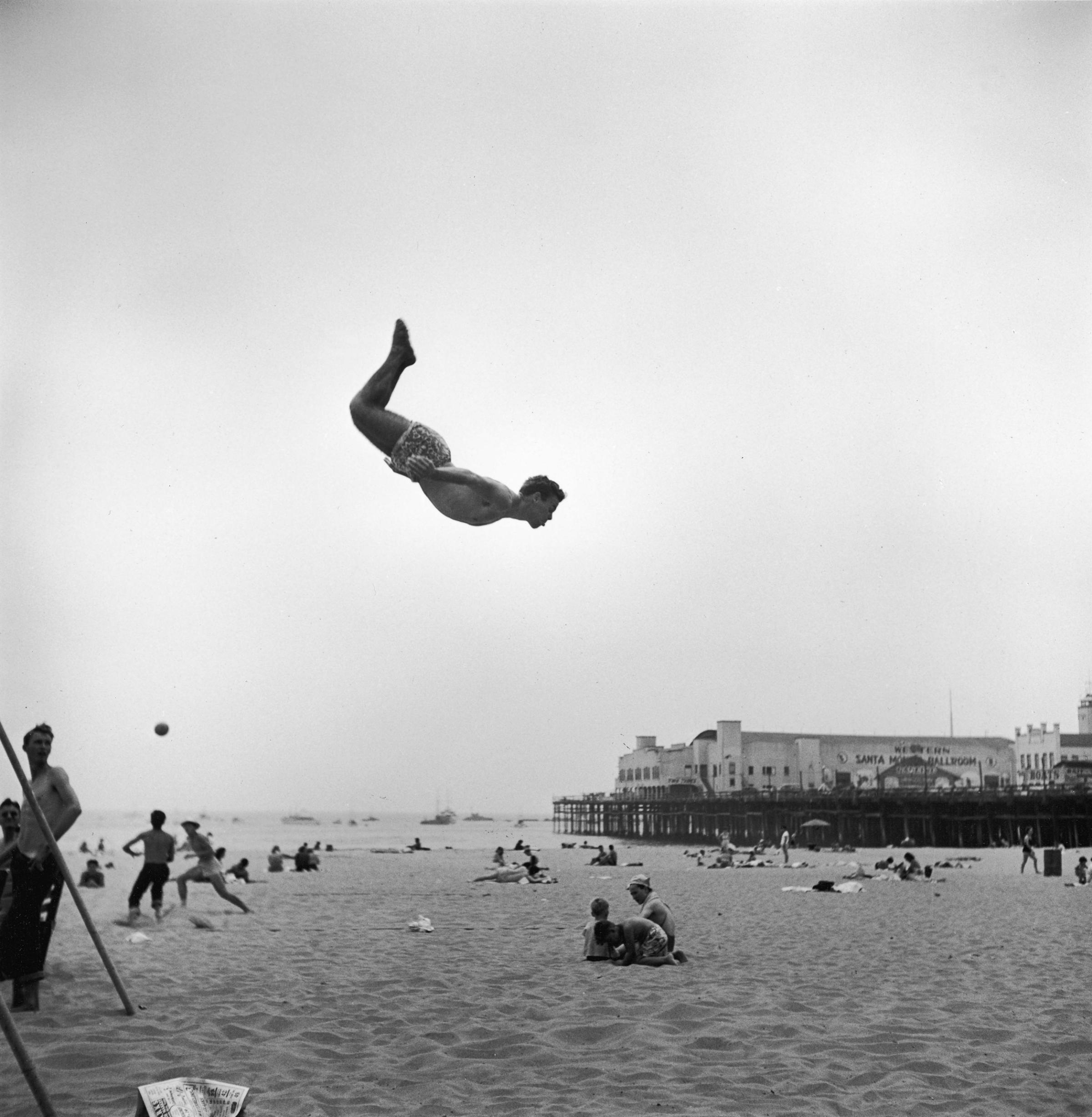 Daring young man flies off trampolin at California's Santa Monica Beach, where movie starlets show off.