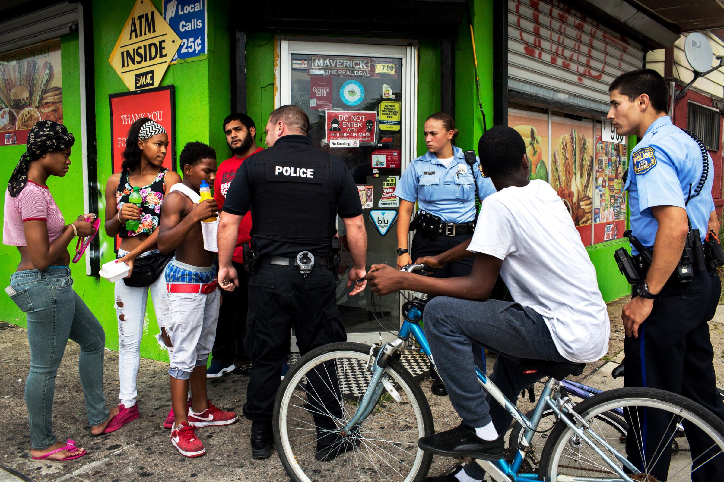 July 28th, 2015. Philadelphia, PA. After a shooting nearby with reports of a suspect fleeing on a bicycle, officer Ryan Mundrick (center, black clothes) joins "foot beats" Ashley Sipos (center right police officer) and Jonathan Dedos (right) in questioning a group of youth as they come out of a bodega. Dispatch had sent out information that there was a black male suspect on a bicycle so police fanned out looking for people of that description, including many of the youth in the neighborhood. After a brief interaction the police and youth went their separate ways without incident. (Natalie Keyssar)