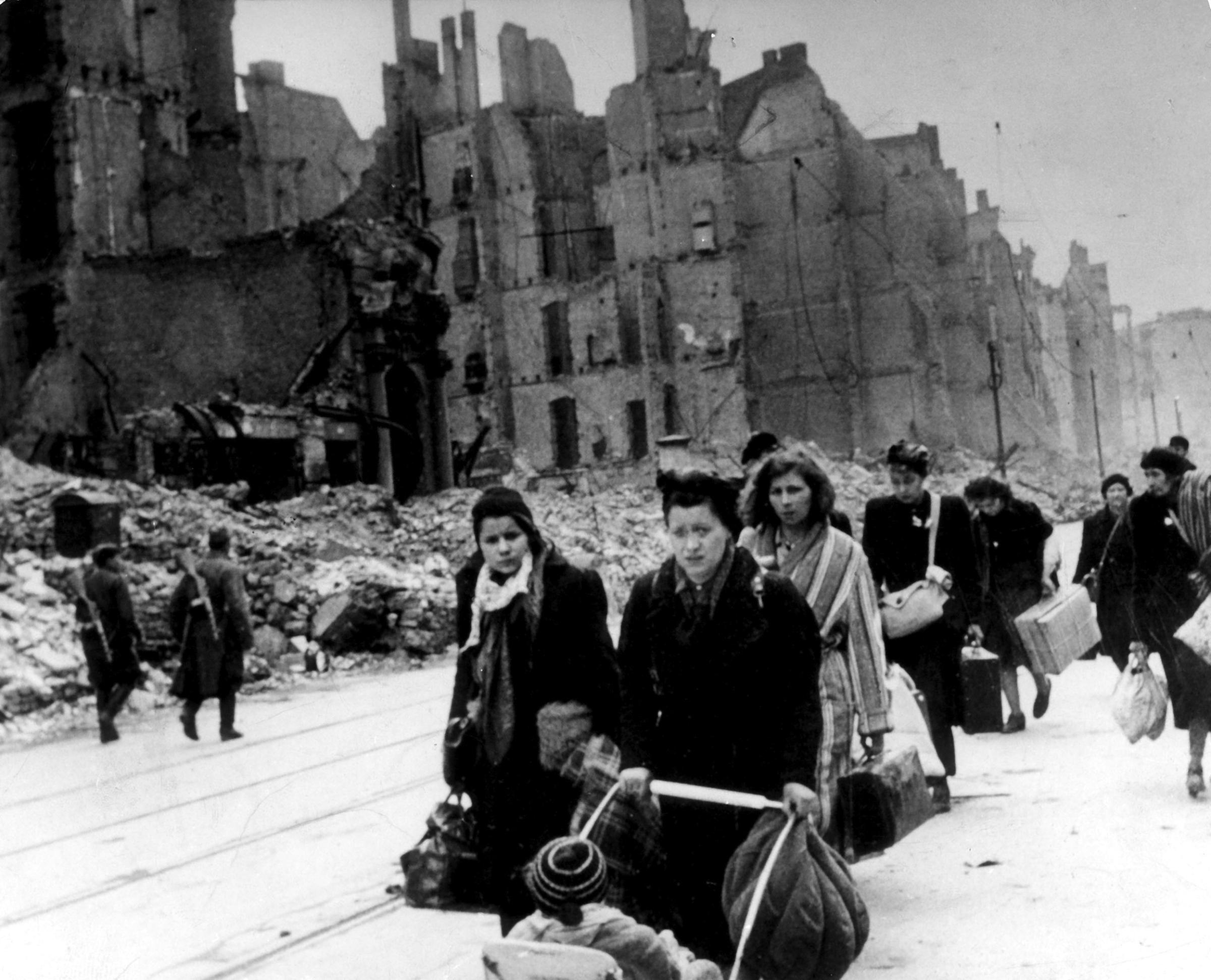 Stream of refugees and people who have been bombed out of their homes moving through destroyed streets - 1945after end of war; on the left two soviet soldiers patrolling).