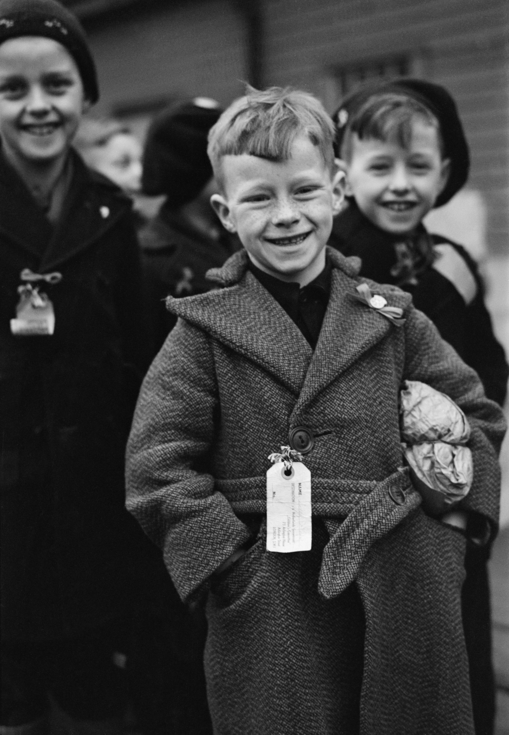 Dutch Child Refugees: Arrival In Britain At Tilbury, Essex, England, UK, 1945, A small Dutch boy smiles for the camera upon arrival at Tilbury in Essex. He is carrying a small paper parcel under his arm, which contains all his luggage. He, and the other children, (some of whom can be seen behind him) all have labels pinned to their coats which bear their names, home address and destination, 11 March 1945.