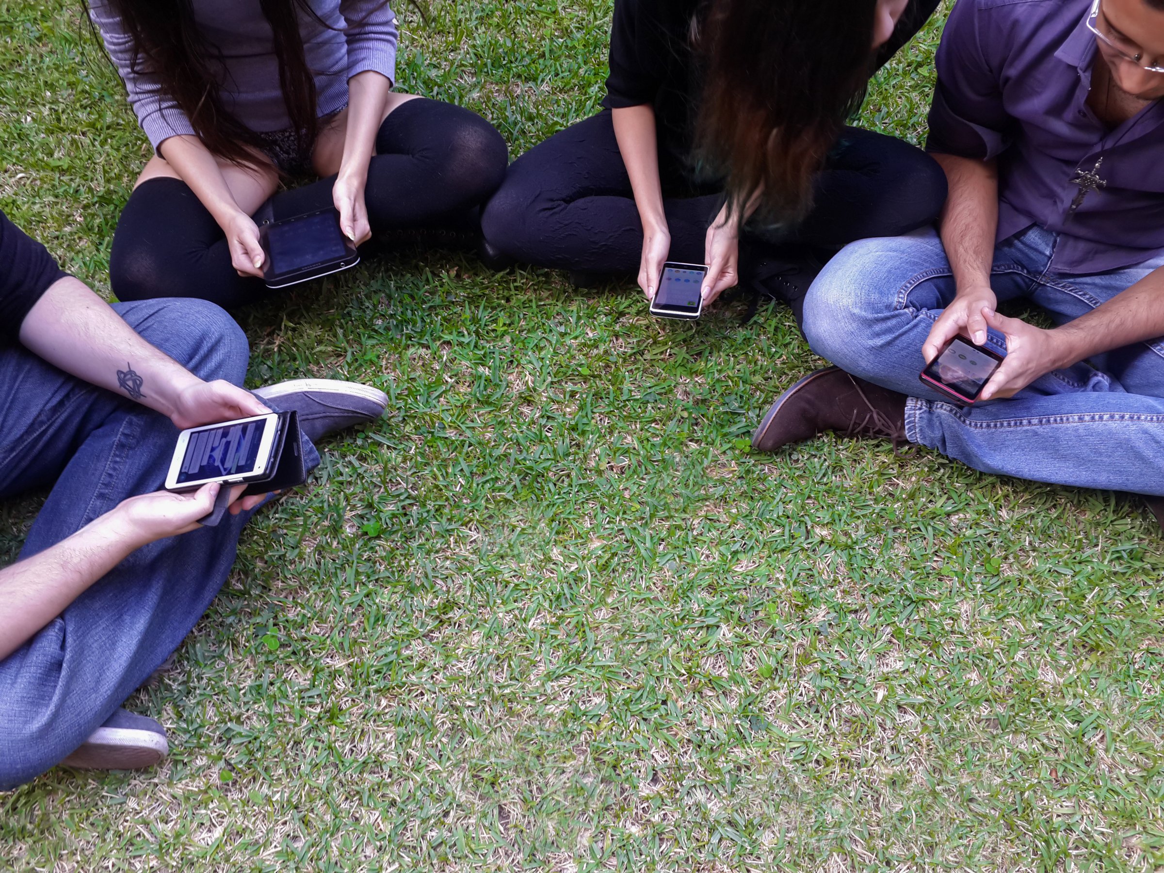 Millennials sit in a circle on grass looking at their phones.