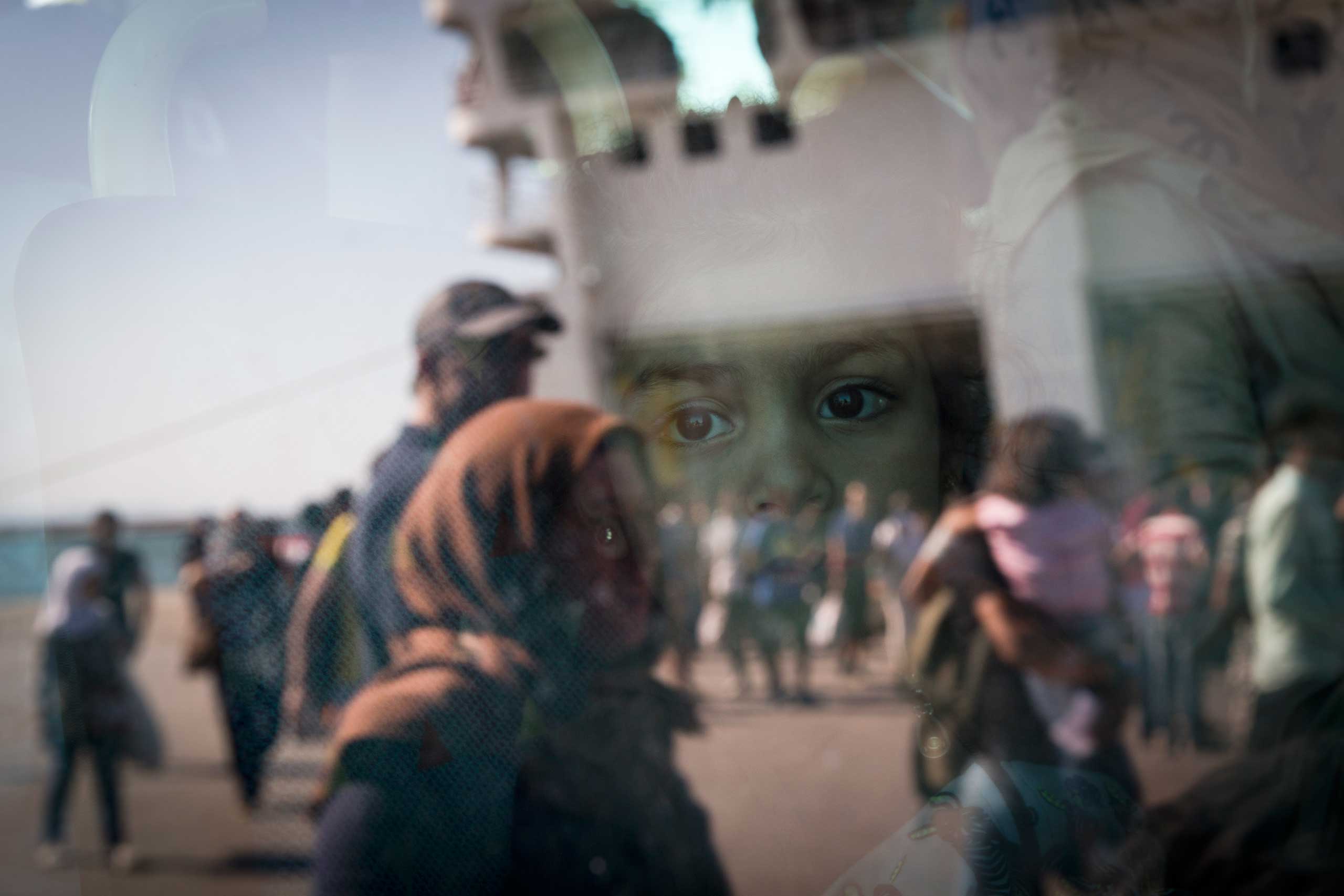 A little girl from Syria looks out of a bus as the ferry she arrived in is reflected in the bus window at the port of Piraeus, Greece, on Aug. 25, 2015.
