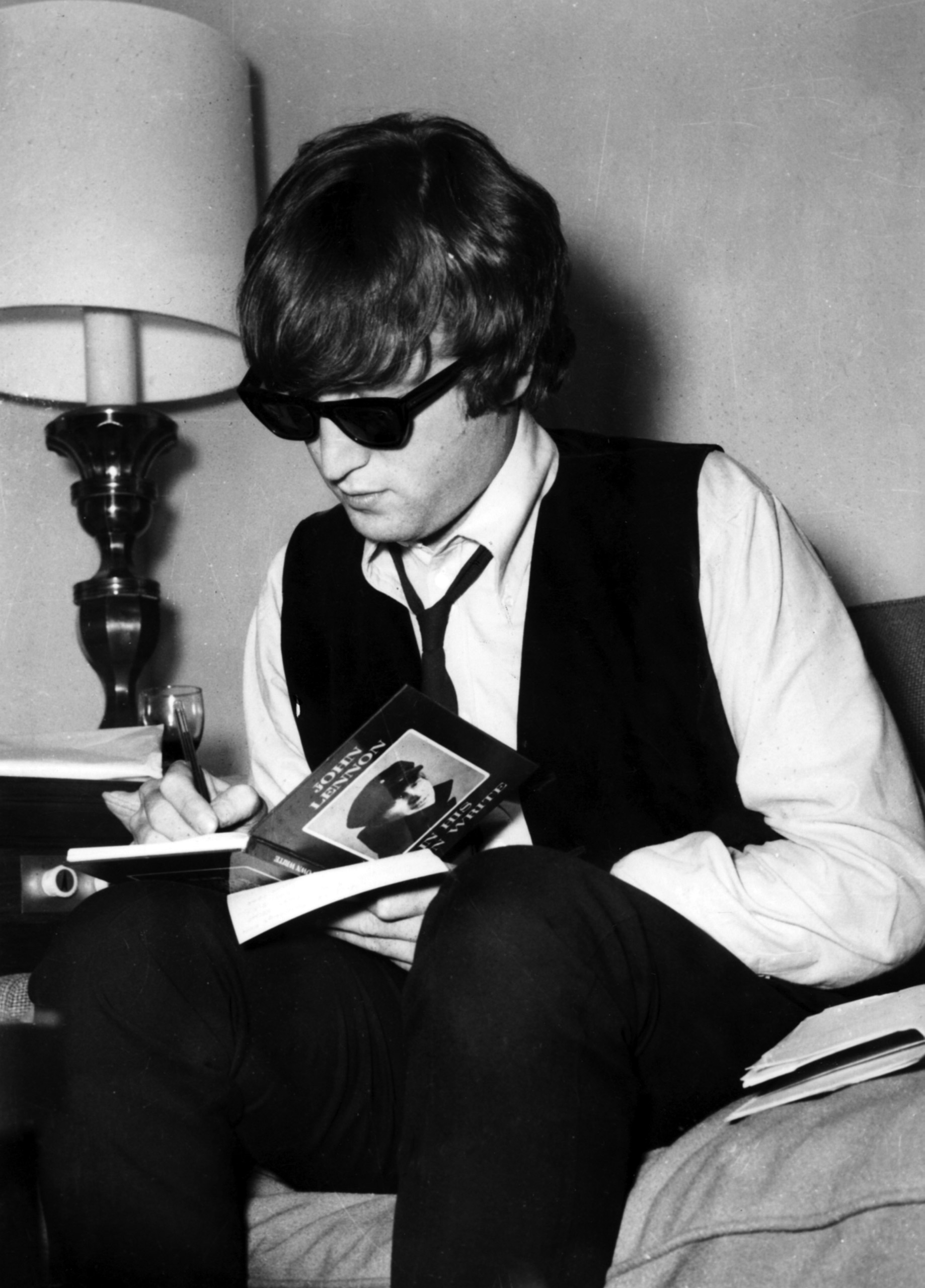 John Lennon sitting on bed in sunglasses signing a book.