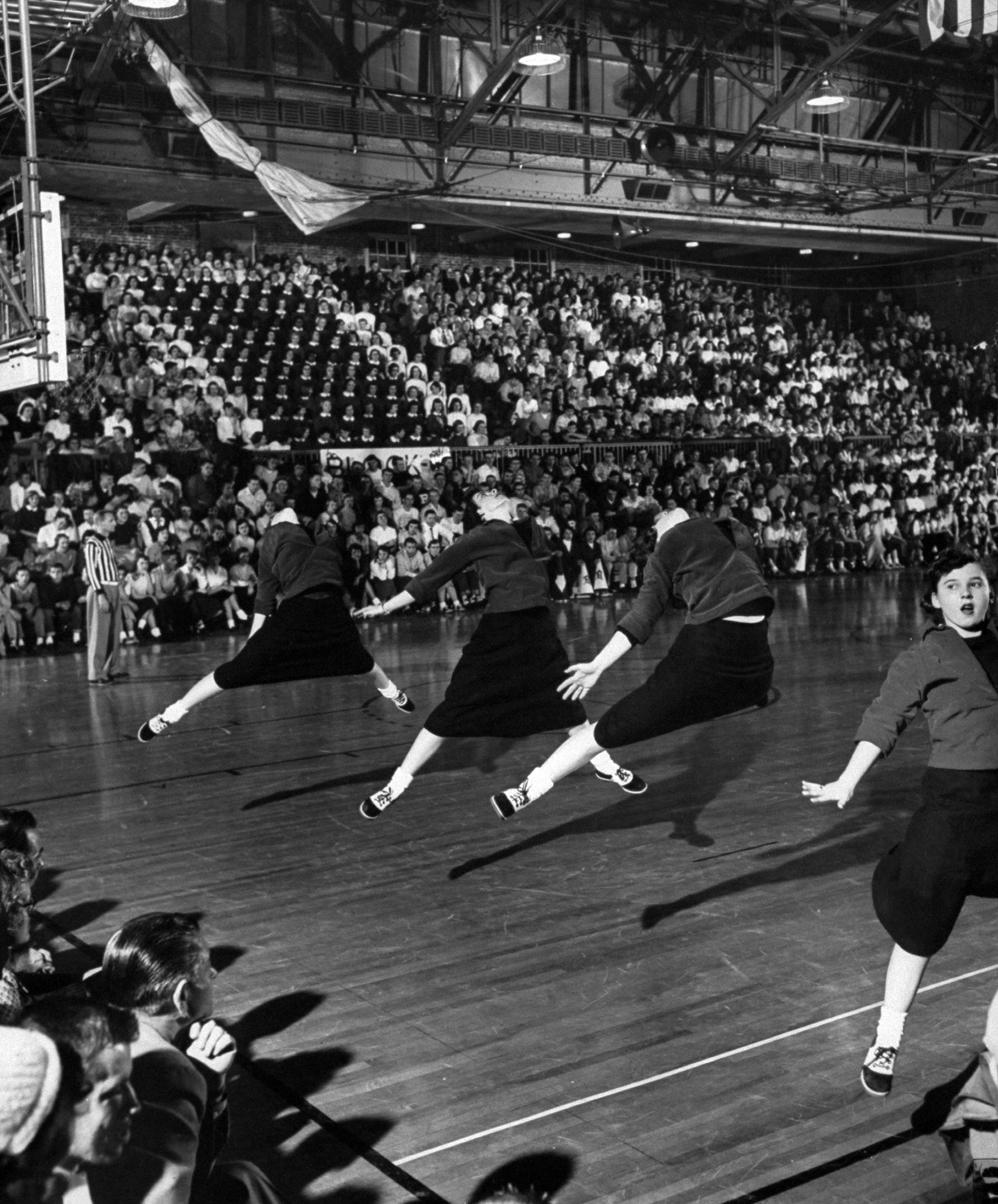 High school girl cheerleaders wearing sweaters and skirts leaping high in the air during their vigorous cheers at the basketball game, 1953.