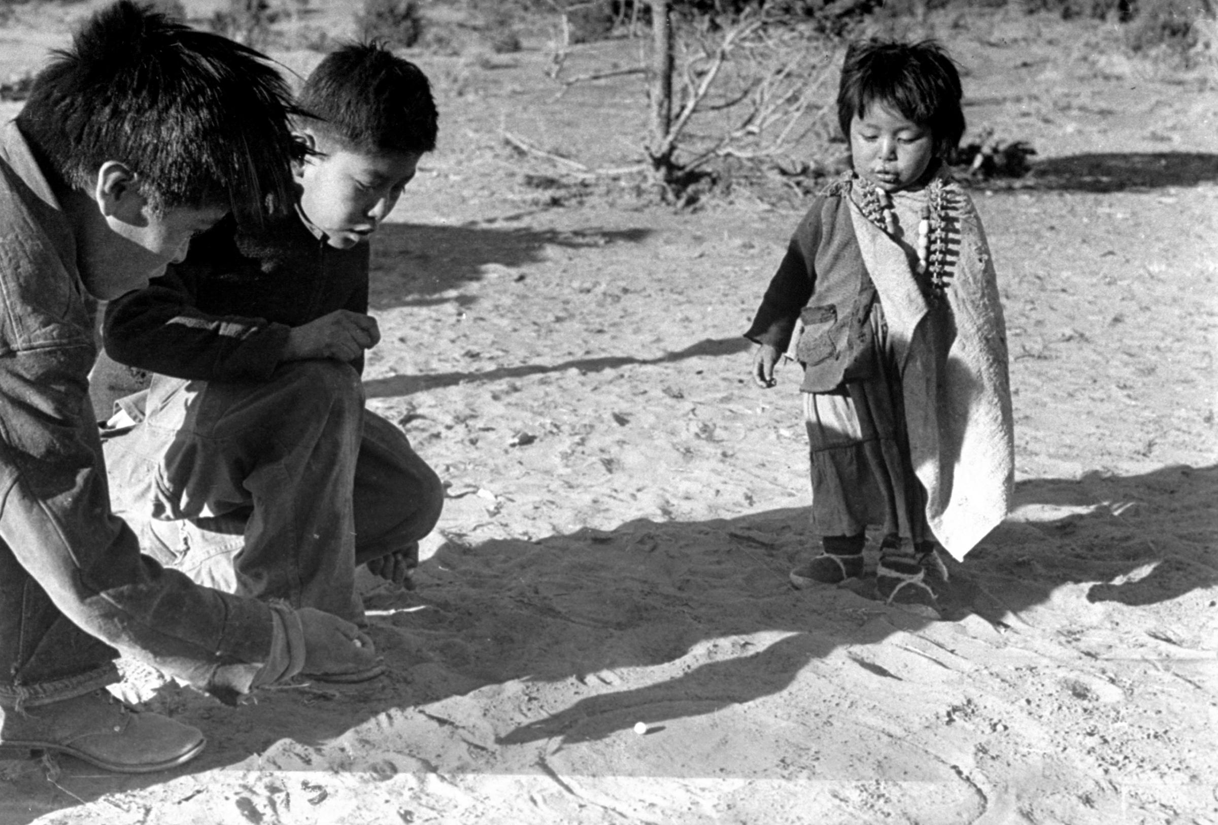 Game of marbles, one popular part of white man's culture, is explained by small boy at center to brother and sister. This boy goes to school and learned the game there. His brother has to stay home to help with the sheepherding.