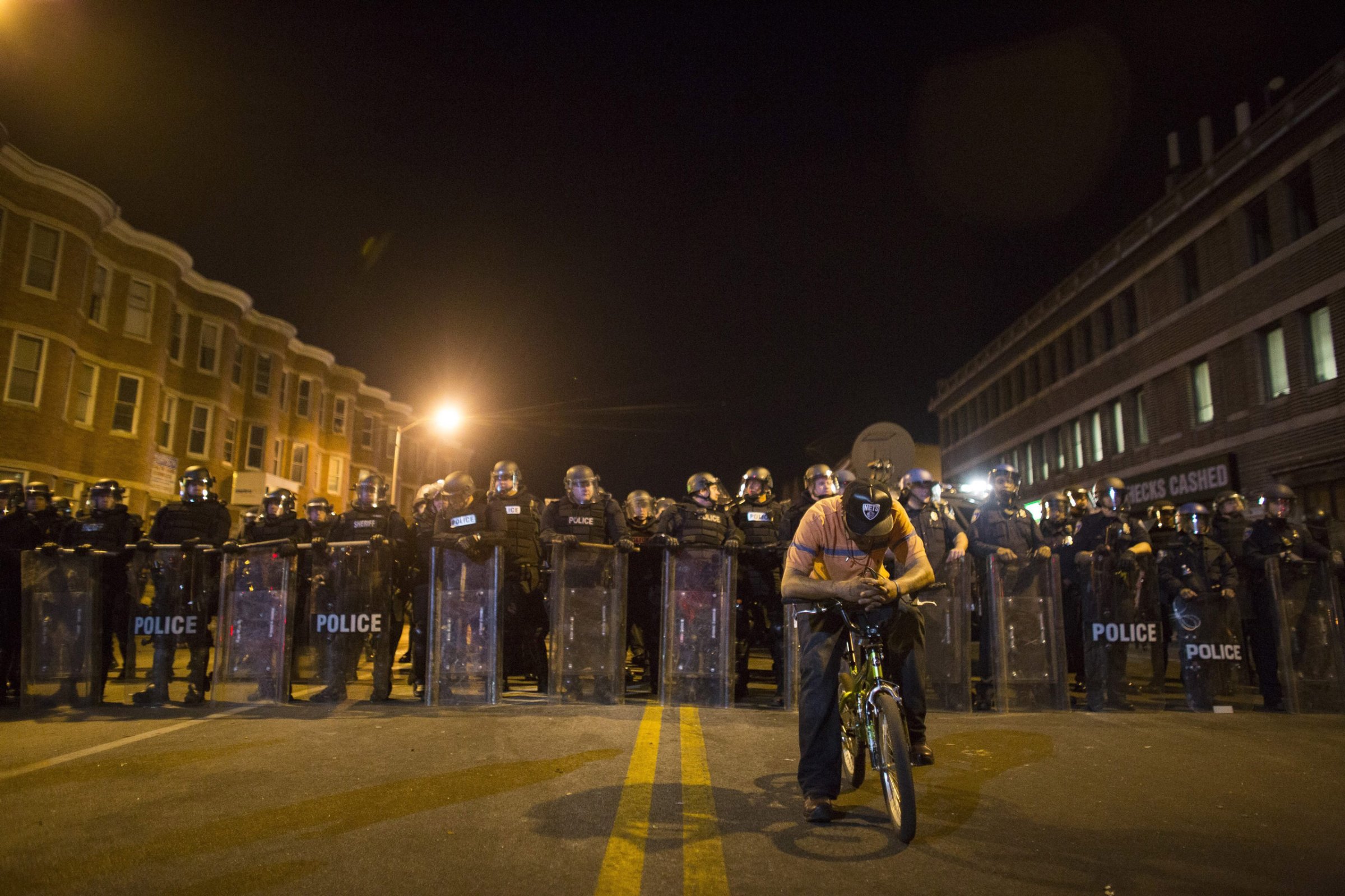 epa04988256 YEARENDER 2015 APRIL A protester on his bike is seen in front of a line of policemen in riot gear during a 10 p.m. curfew in Baltimore, Maryland, USA, 28 April 2015. Tensions eased on 28 April after peace-keeping civilians kept rock-throwing protestors at bay from lines of police in riot gear. EPA/JOHN TAGGART