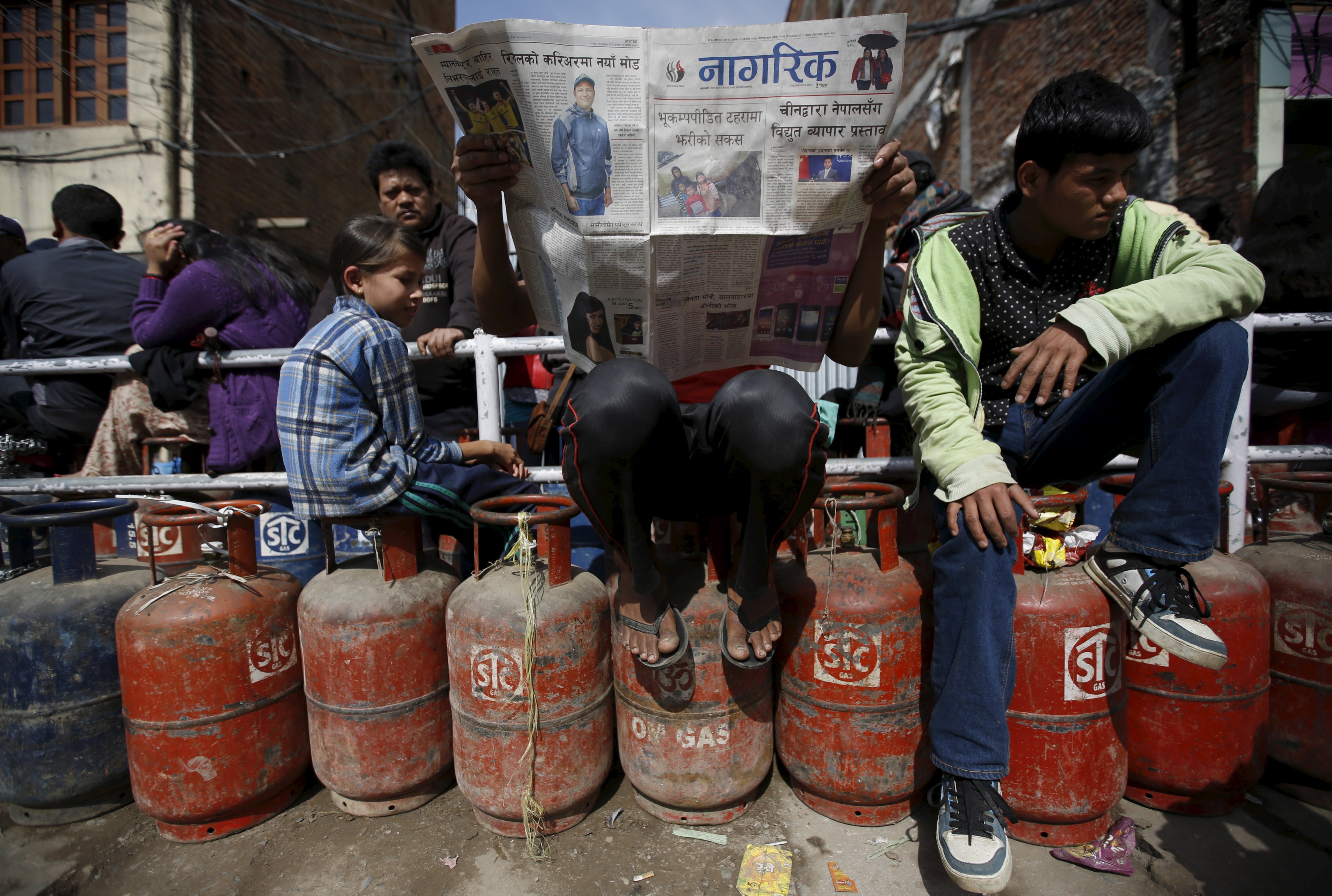 A man reads a newspaper as he sits on top of empty gas cylinder as he waits in a queue to buy cooking gas along with other customers during the ongoing fuel crises that has been continuing for over a month now in Kathmandu