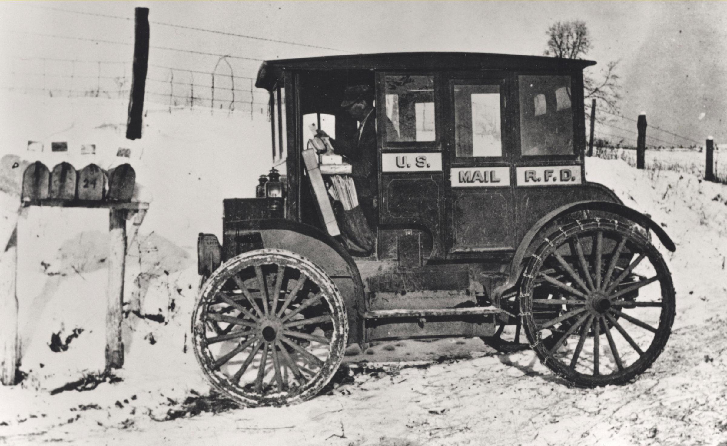 Postal officials encouraged Rural Free Delivery (RFD) carriers to replace their horses and wagons with the latest in transportation technology. This unidentified carrier painted his early electric-motored vehicle in the same paint and identification scheme as the RFD wagons of the era. He is, no doubt, only able to complete his wintertime rounds thanks to a snow-plowed road. Automobiles were not yet adequate replacements for horses, wagons, and sleds on rural roads. 1910.