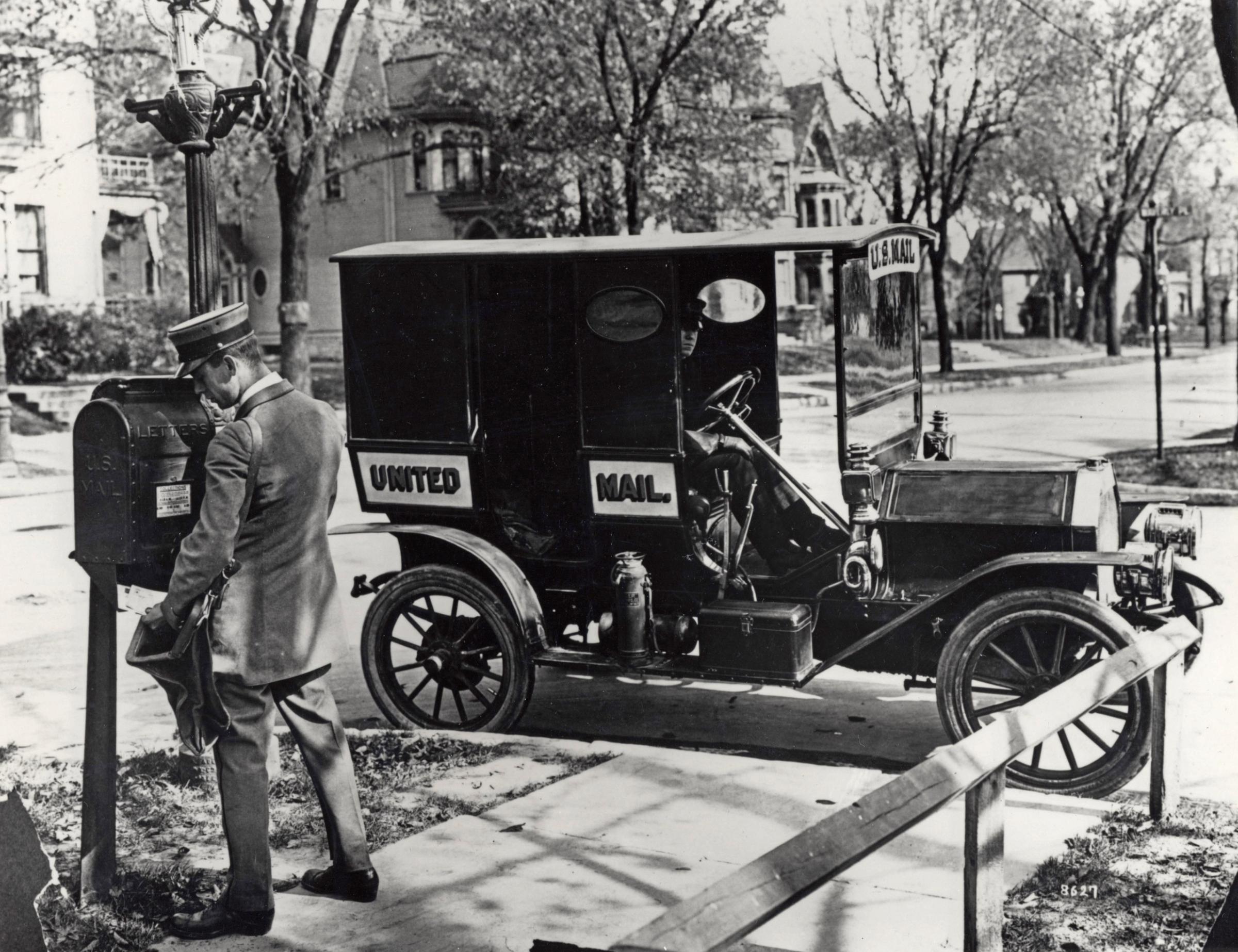 A letter carrier collecting mail from a sidewalk mail collection box. His mail truck is parked at the curb. 1915.