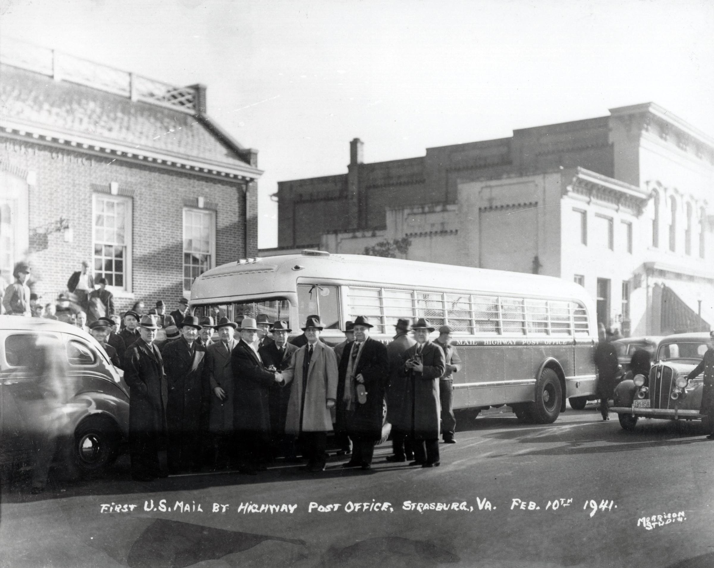 Officials are gathered to welcome the first Highway Post Office bus in Strasburg, Virginia on February 10, 1941. This bus traveled on a route between Washington, DC and Harrisonburg, Virginia. By the 1930s, a significant decline in railroad passenger traffic had caused a subsequent decline in the use of railway trains. To fill the void, the postal service transferred some en route distribution from trains to highway buses. This is the first Highway Post Office bus and is in the collection of the National Postal Museum. 1941.
