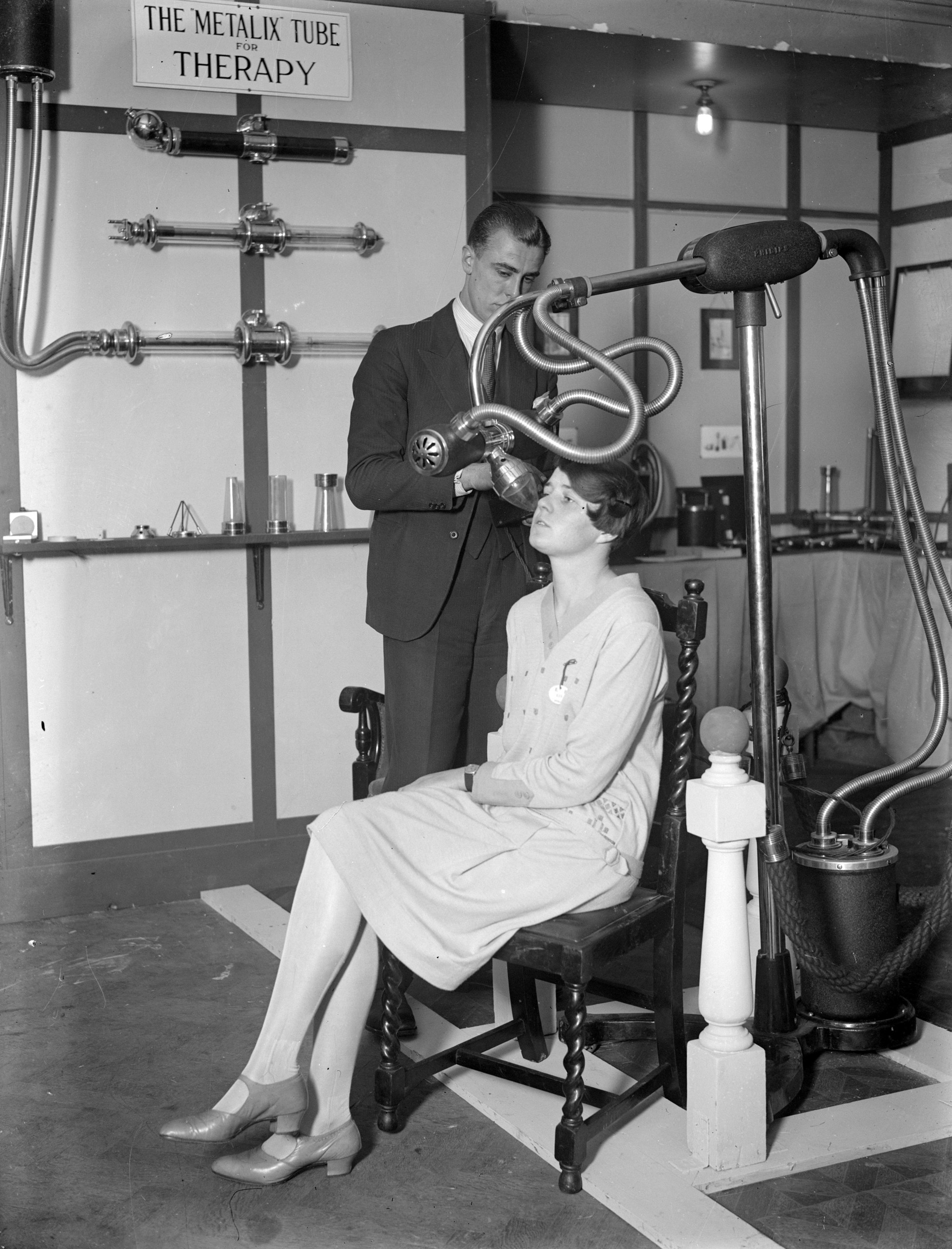 A man and a woman demonstrating medical equipment at a X-ray exhibition, beside a sign reading 'The Metalix Tube for Therapy,' 1928.