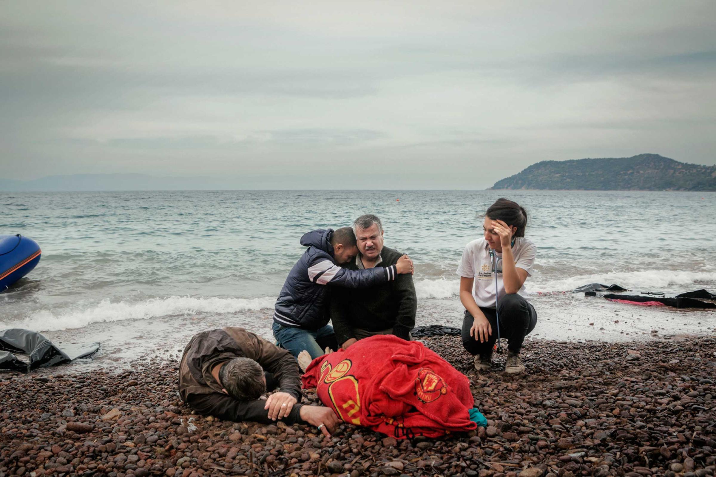The husband (right) and relative of a 65-year old Iraqi refugee woman grieved as her body is covered with a towel on the Greek island of Lesbos, Greece, Oct. 16, 2015. A man who made the journey with them and a volunteer stood nearby. The woman reportedly drowned after smugglers violently forced the family to leave the Turkish coast in an inflatable boat, which immediately began to sink after they departed.