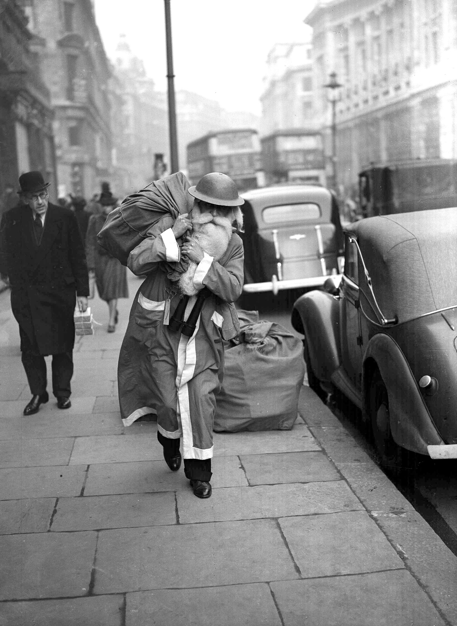 Father Christmas walks the streets of wartime London, The old man has exchanged his civilian red hood for a warlike 'tin hat', but, blitz or no blitz, he is delivering goods this year. December 1940.