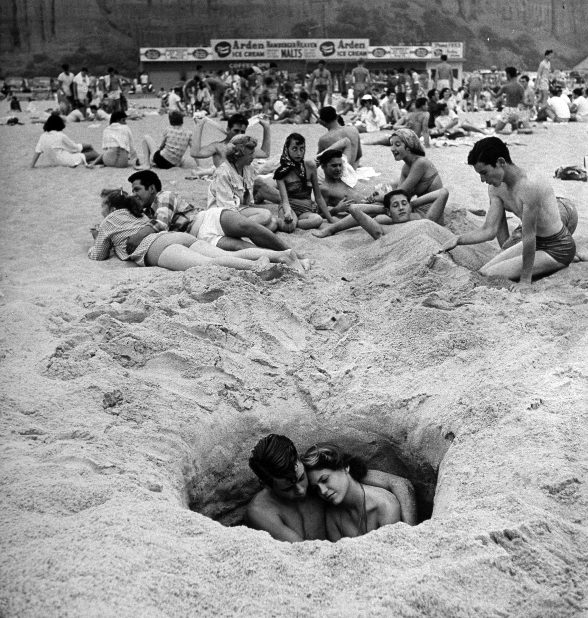 Young couple cuddling as they sit down in a hole in the sand while others lie around behind them on a hot Independence Day at the beach, 1950.