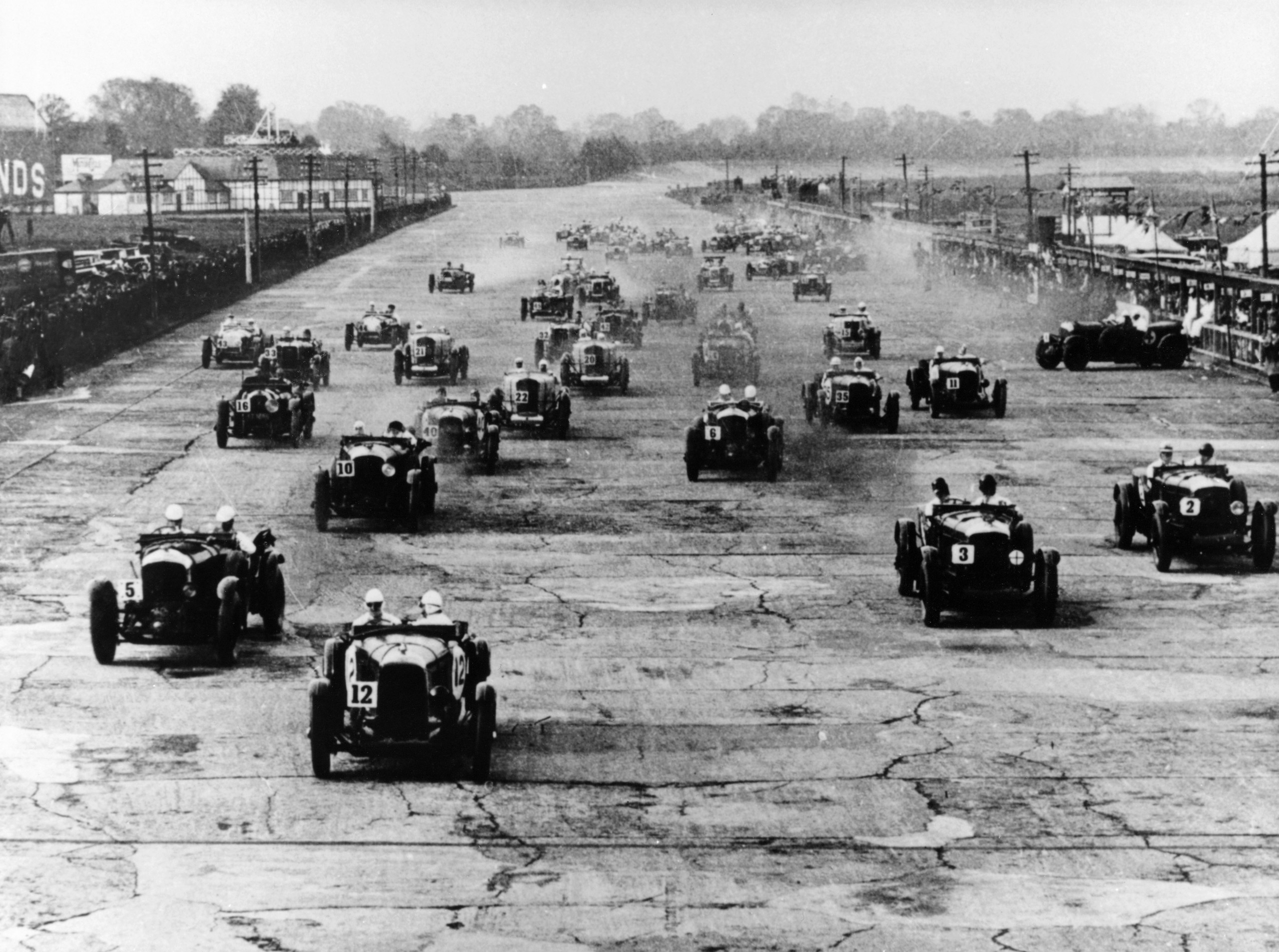 Motor race, Brooklands, Surrey, 1920s. A large field closely bunched together. On the right, one of the competitors appears to have spun.