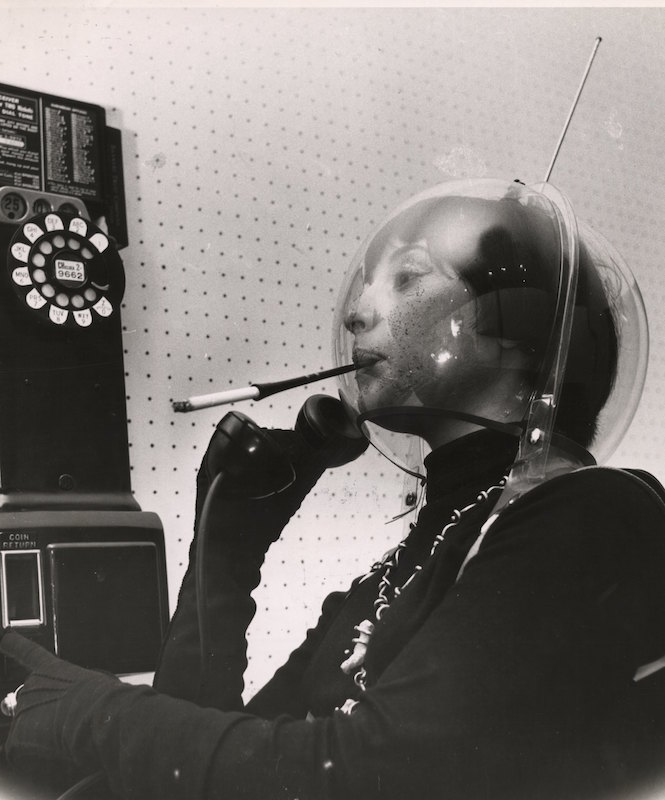A young woman wears a plastic globe on her head, like an alien or an astronaut, smoking a cigarette in a holder and holding the receiver of a pay telephone, in Greenwich Village, New York, New York, mid 1950s. (Weegee(Arthur Fellig)—International Center of Photography / Getty Images)