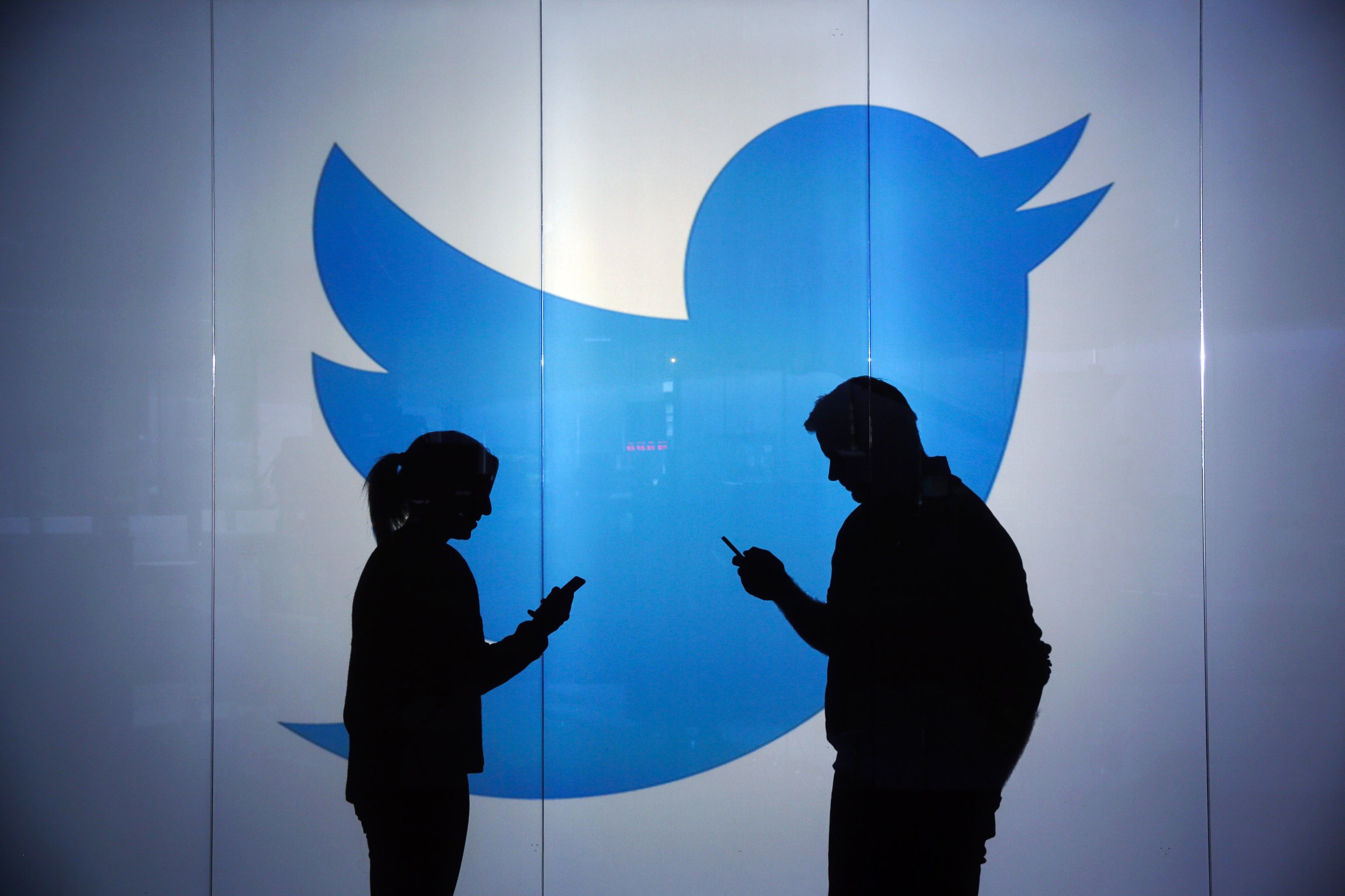 People are seen as silhouettes as they check mobile devices whilst standing against an illuminated wall bearing Twitter Inc.'s logo in London, U.K., Jan. 5, 2016.