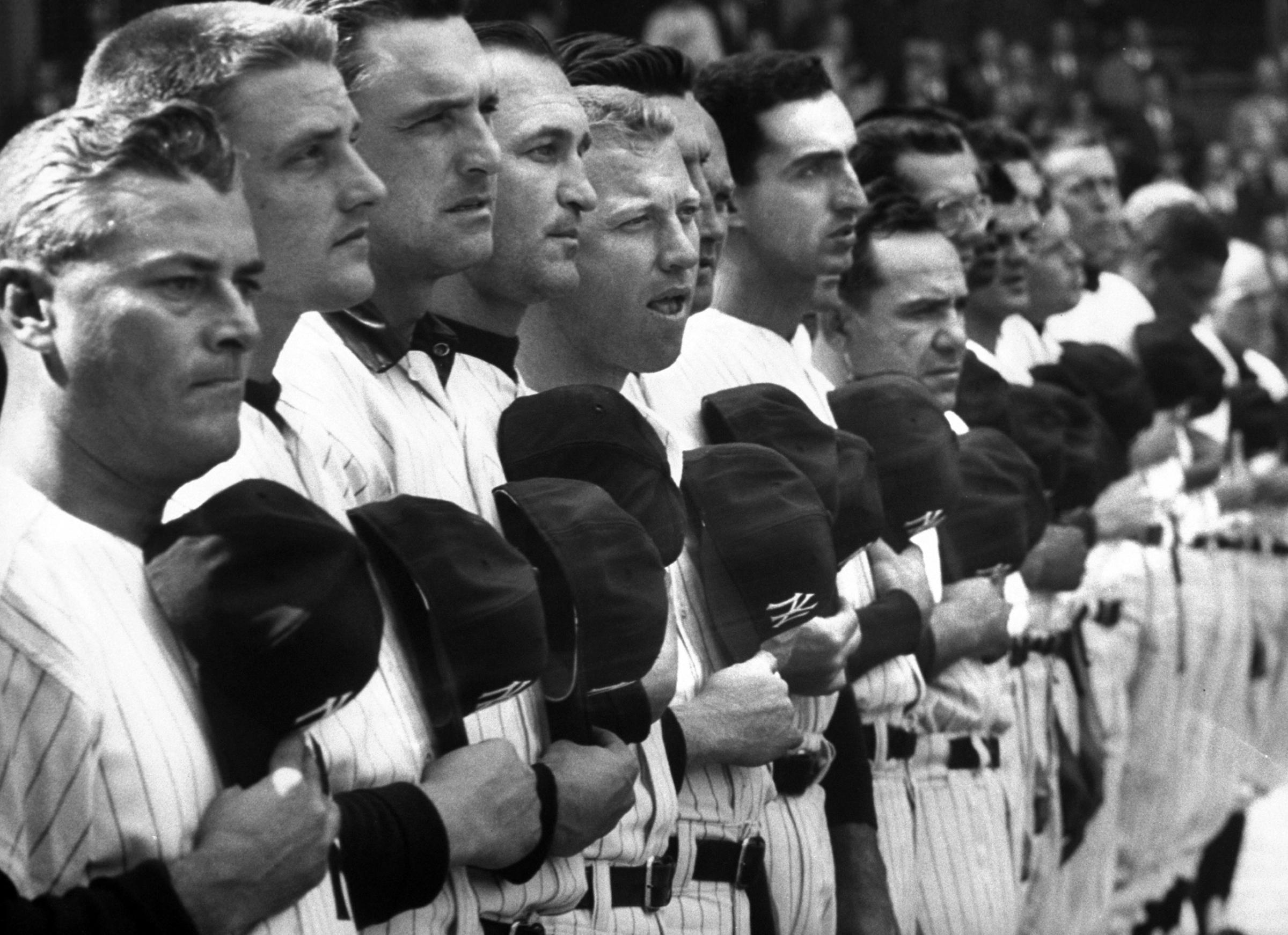 NY Yankees Roger Maris (2L), Mickey Mantle (5L) &amp; Yogi Berra (9L) standing in line with other Yankees holding baseball caps over hear during playing of National Anthem on opening day. 1962.