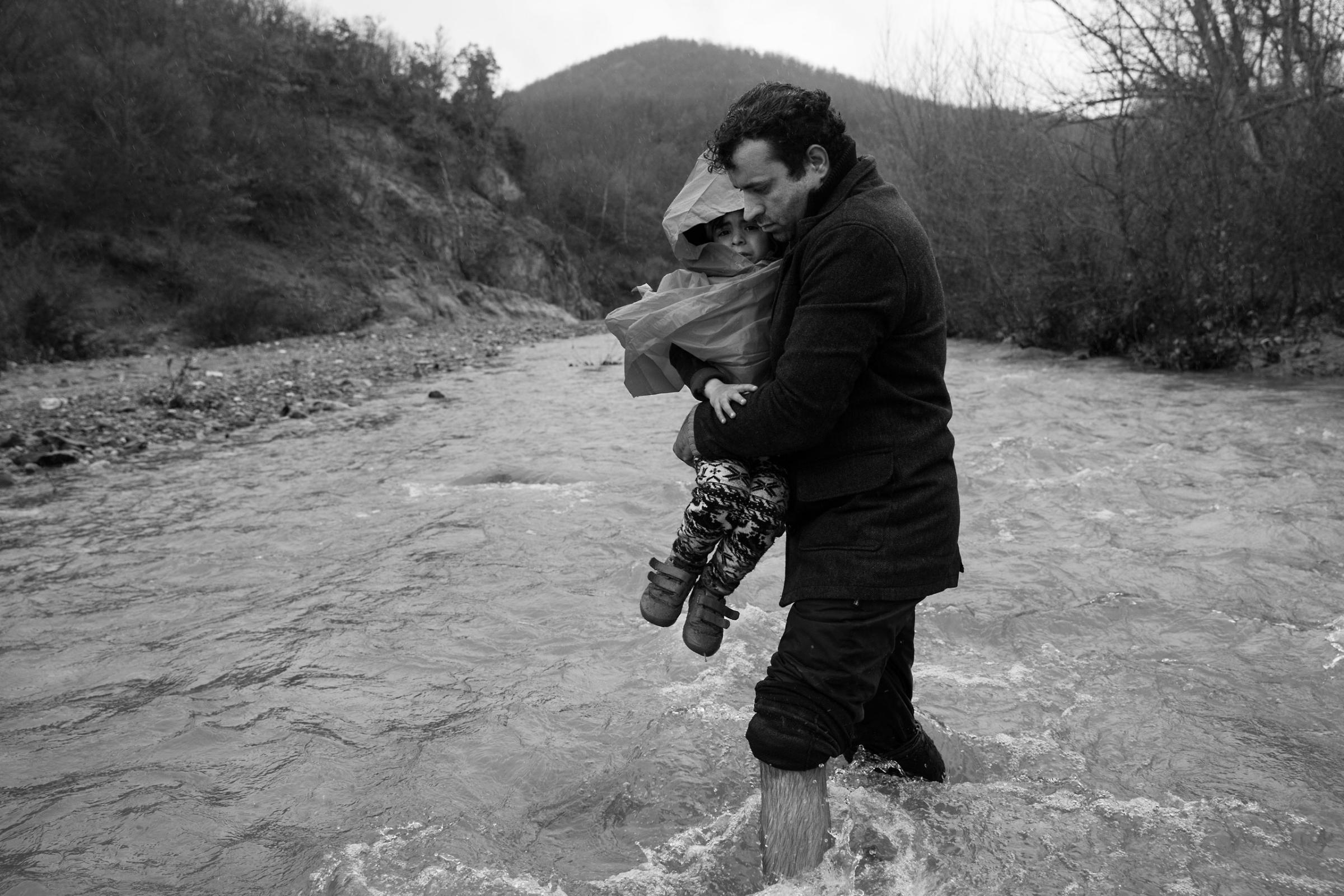 A man carries his child across a river into Macedonia on March 15, 2016 Scores of refugees, including elderly, disabled, and families with children attempted to make the dangerous trek.