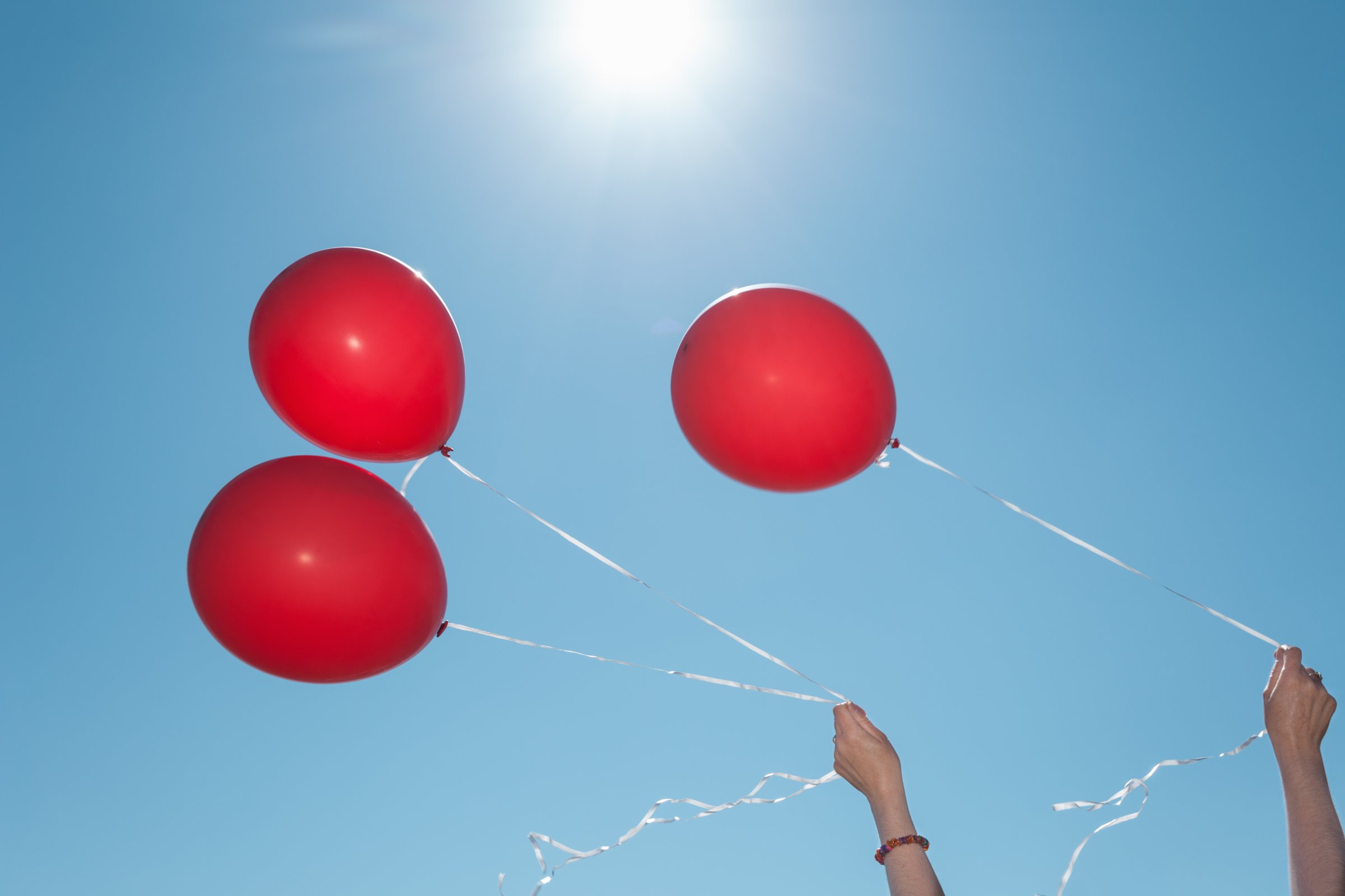 Hands holding three red balloons against blue sky