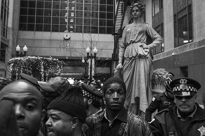 Black Lives Matter protestors in front of the Chicago Board of Trade during the LaQuan McDonald protests. Hundreds of people continue to protest the ongoing aftermath of police killings of black men in the City of Chicago. The most recent video to be released was that of 25 year old Ronald Johnson. He was shot and killed by Chicago Police while resisting arrest and running away in 2014. Police and state prosecutors claim that the video shows a gun in Johnson's hand as he ran away and was shot in the back, but activists and family membesr dispute the fact. Charges will not be filed against the officer who killed Johnson. Additionally, 17 year old Laquan McDonald was shot and killed at the hands of Chicago police officer Jason Van Dyke. McDonald was shot sixteen times and the video was withheld from public view until a judge ordered it released. McDonald was holding a small knife but was shot repeatedly in the dash cam video. The family was paid 5 million dollars in a settlement with the City of Chicago and recently officer Van Dyke was also charged with murder after a whistleblower brought the video to the attention of a local journalist who then sued the city to have it released. The City of Chicago's Police Department has a long and difficult history associated with police brutality reaching back decades. Protests continue to grow and have led to the resignation of police superintendent Garry McCarthy as well as calls for the resignation of Mayor Rahm Emmanuel. Additionally, the Justice Department has opened an ongoing investigation of the Chicago Police Department's handling of these cases and their use of excessive force.