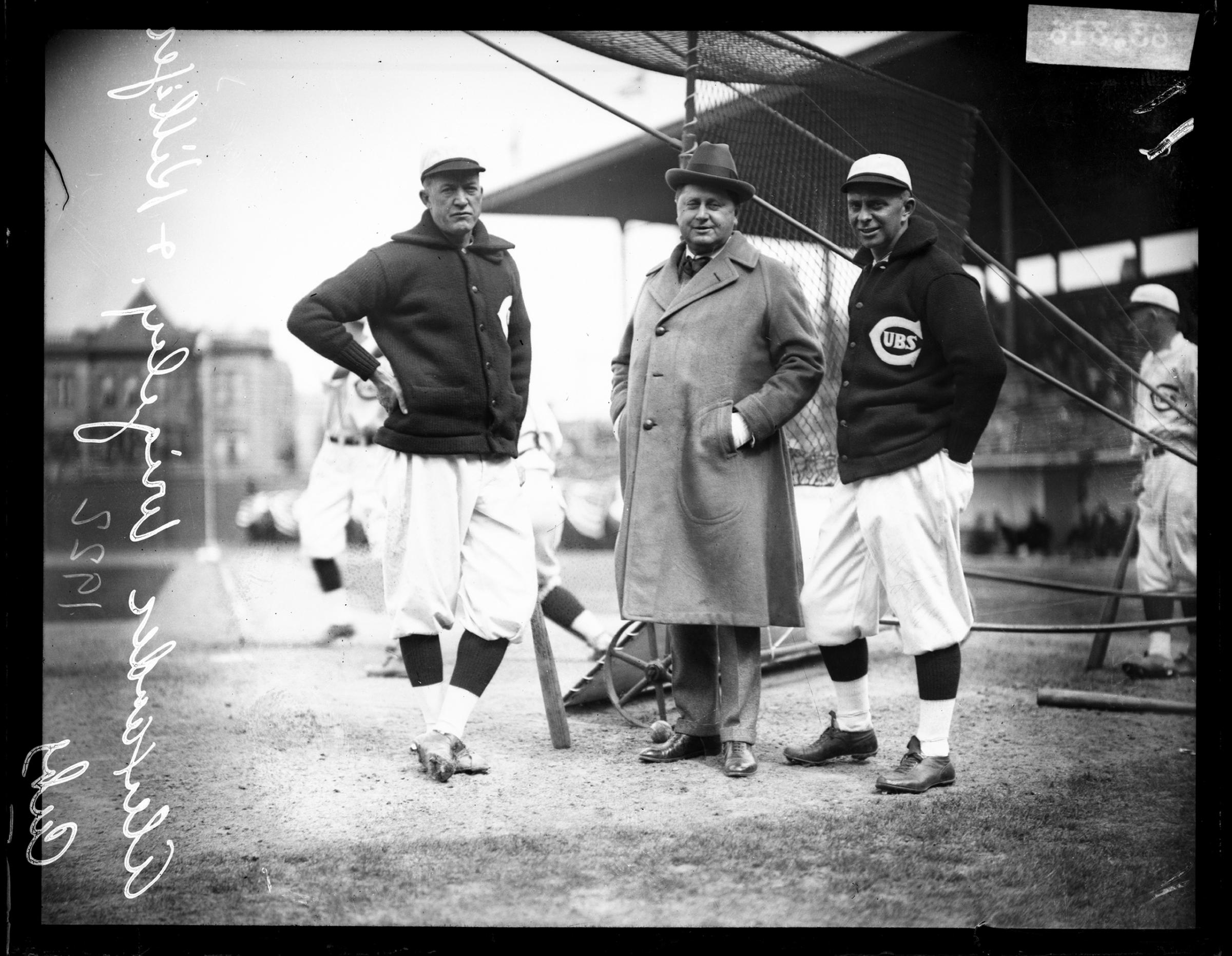 Chicago Cubs baseball player Alexander, co-owner William Wrigley Jr, and manager Bill Killefer standing behind a batting practice backstop on the field at Weeghman Park, located at 1060 West Addison Street, Chicago, Illinois, 1922.