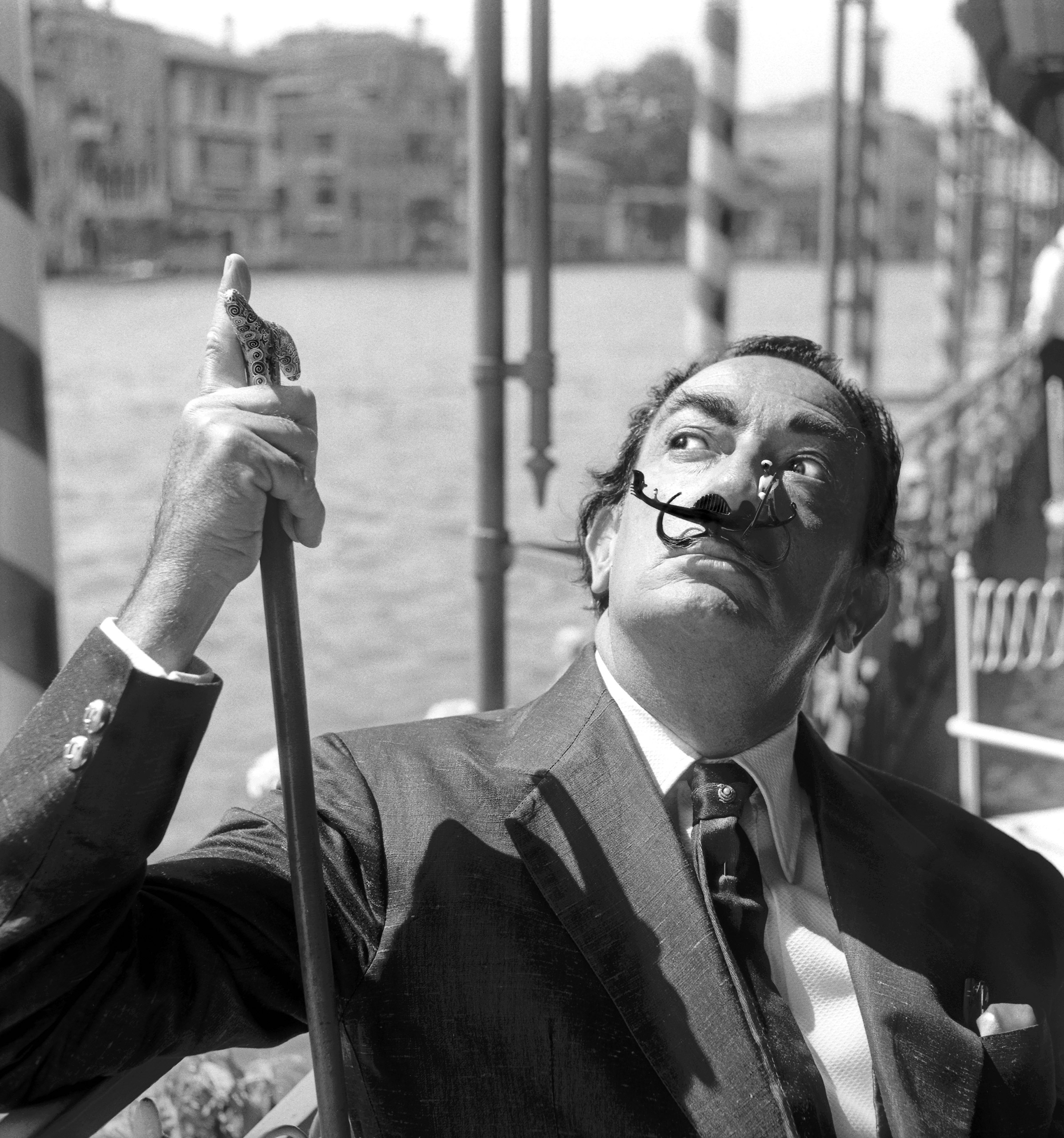 Salvador Dali, wearing a shantung blazer and a silk tie, holdind a walking-stick, with a small toy gondola as a moustache, Venice 1961.