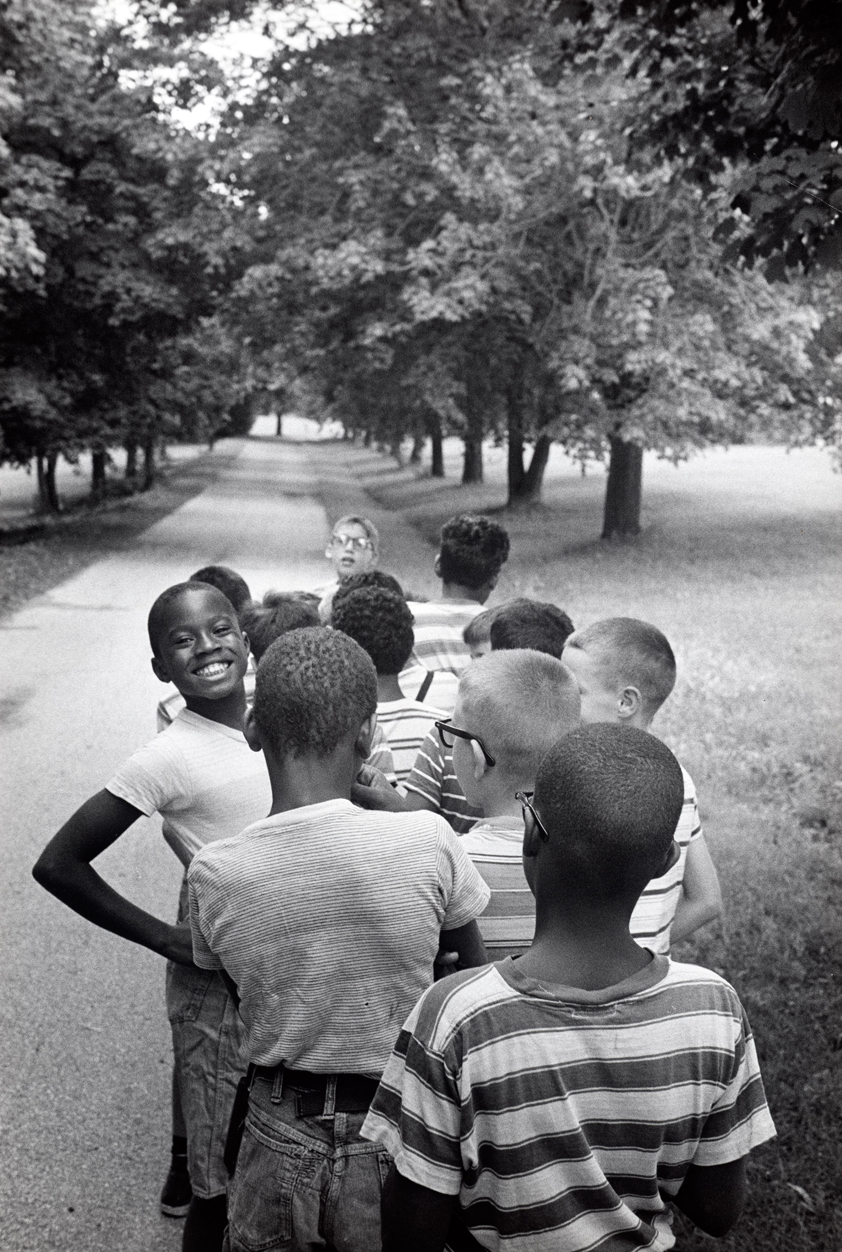 Group of twelve children shown clustered on a tree-lined paved surface. Upstate New York, 1963.