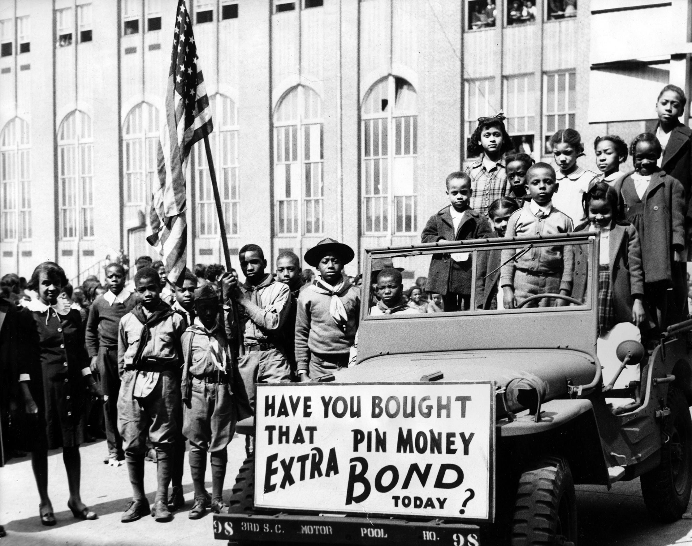 A group of boy scouts carrying the American flag and other children standing in and around a car with a board reading 'Have you bought that pin money extra bond today?, March 24, 1945.