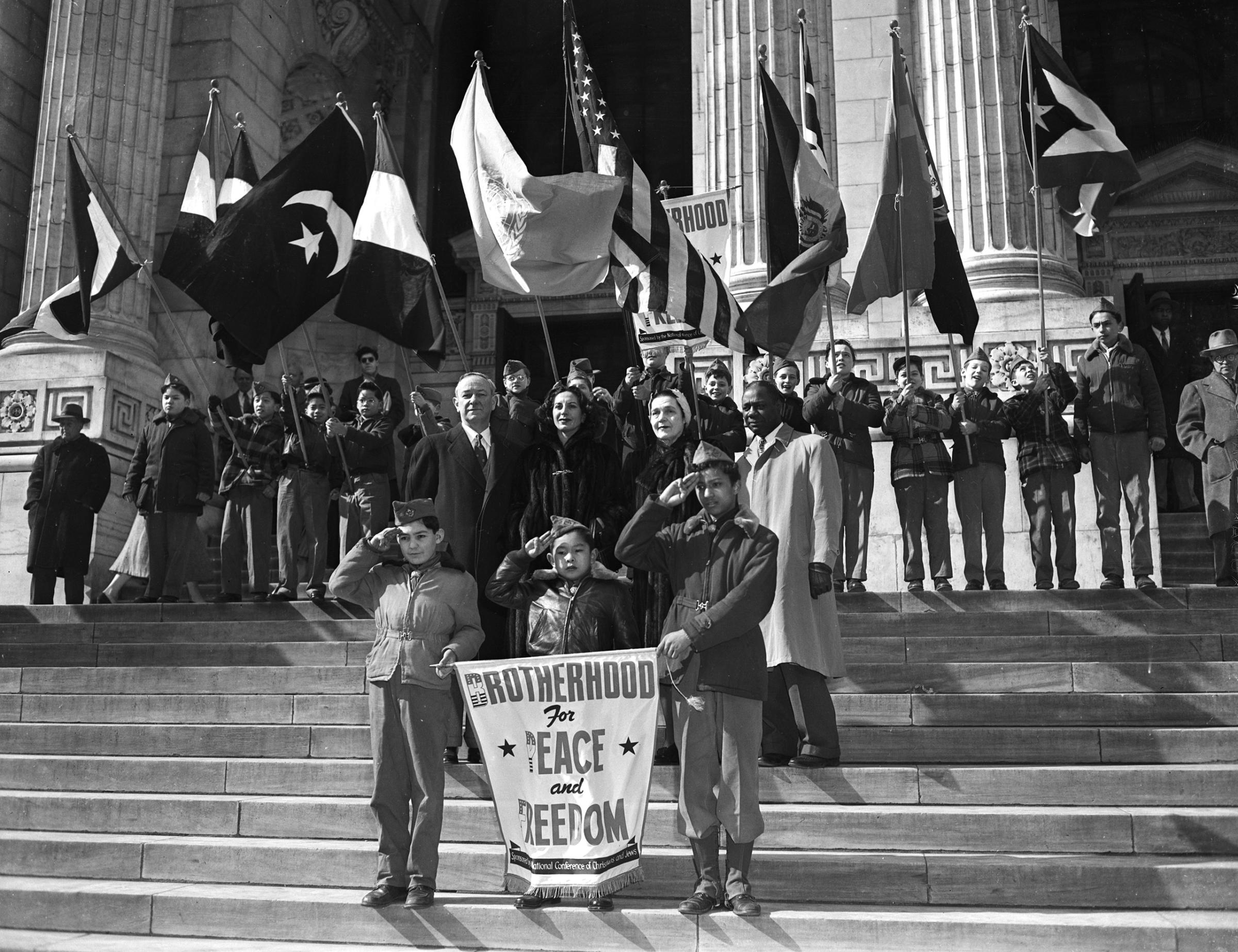 Boy Scouts of various Manhattan troops salute with brotherhood week banner in front of a public library. 1958.
