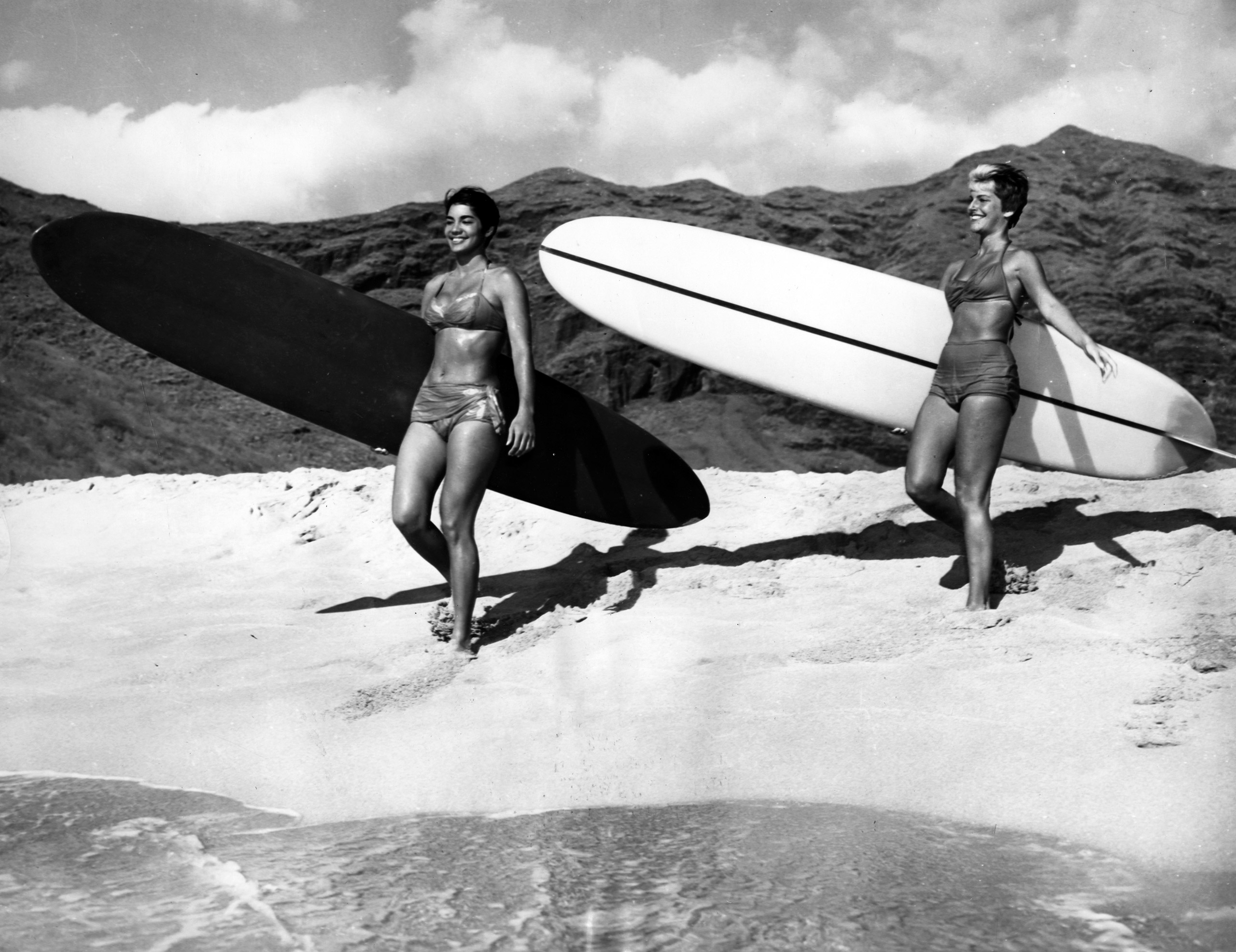 Two women carrying their surfboards at the beach, 1960.